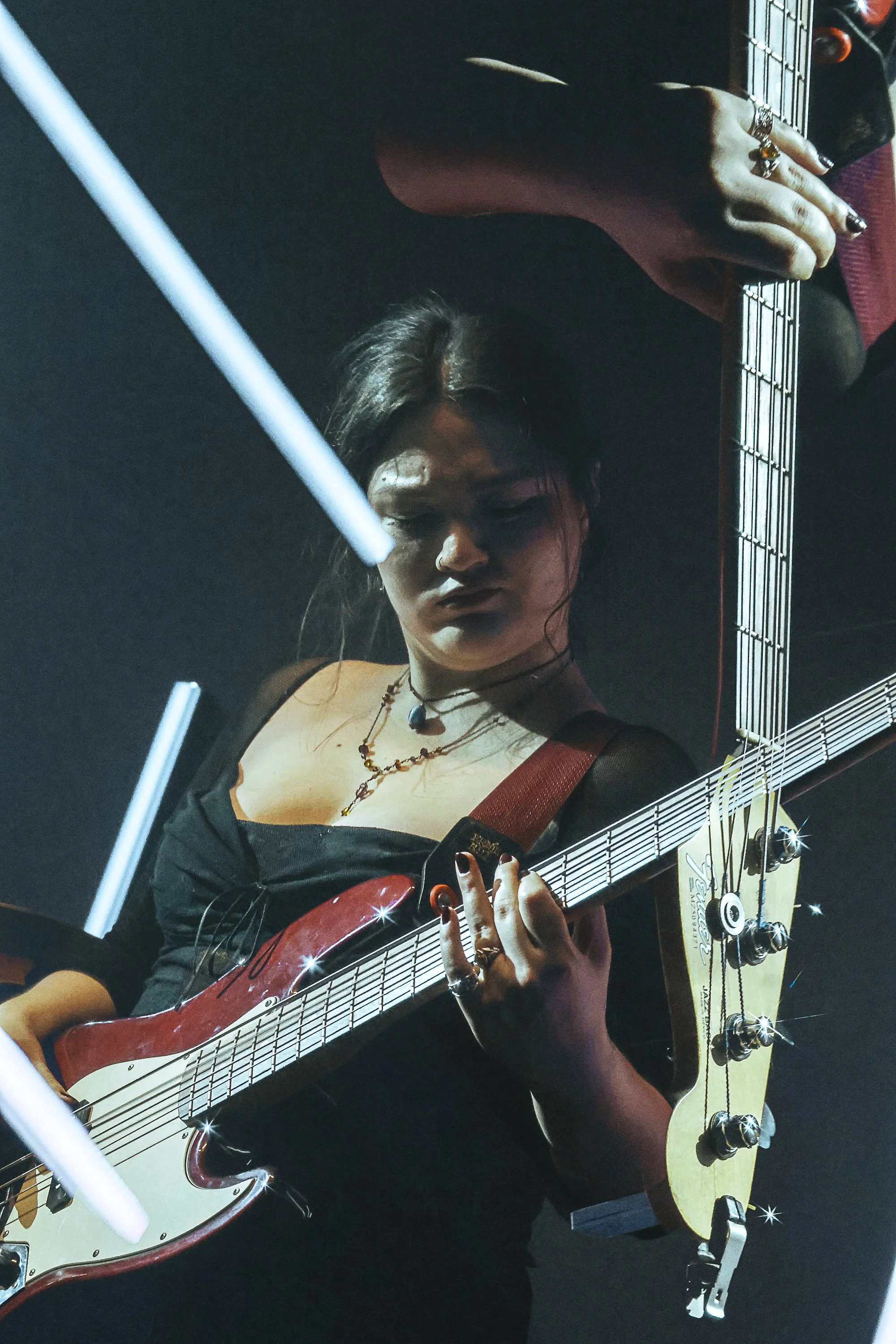 Person playing electric guitar with multiple necklaces, dark hair, and rings, with a second acoustic guitar visible. Purple clothing and blurry background.