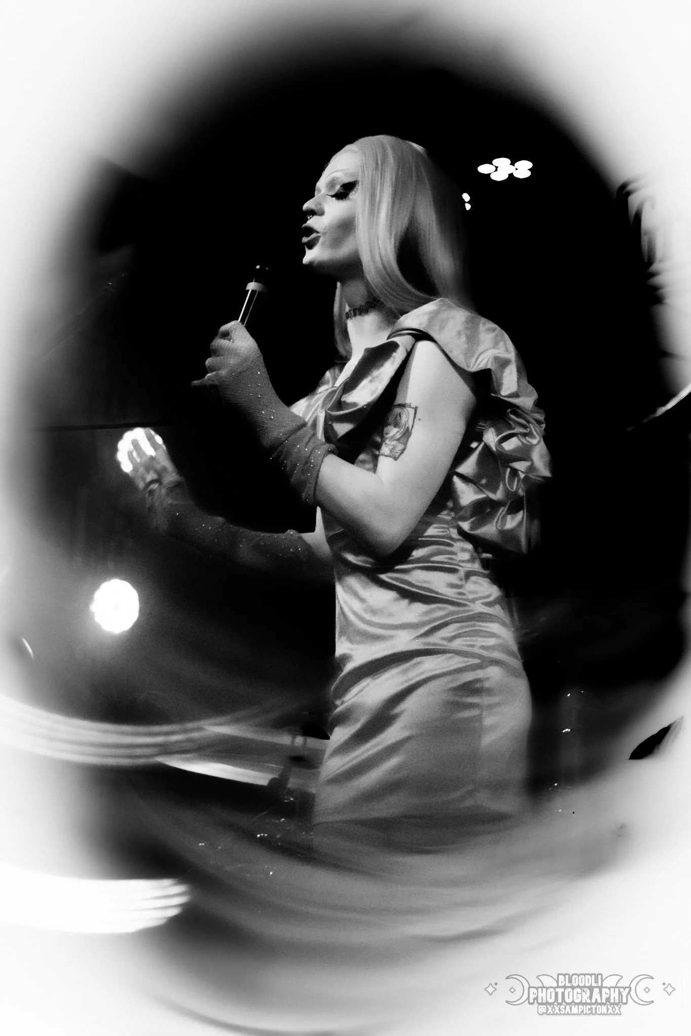 A woman with long blonde hair singing into a microphone on stage, wearing a satin dress and long gloves, captured in black and white.
