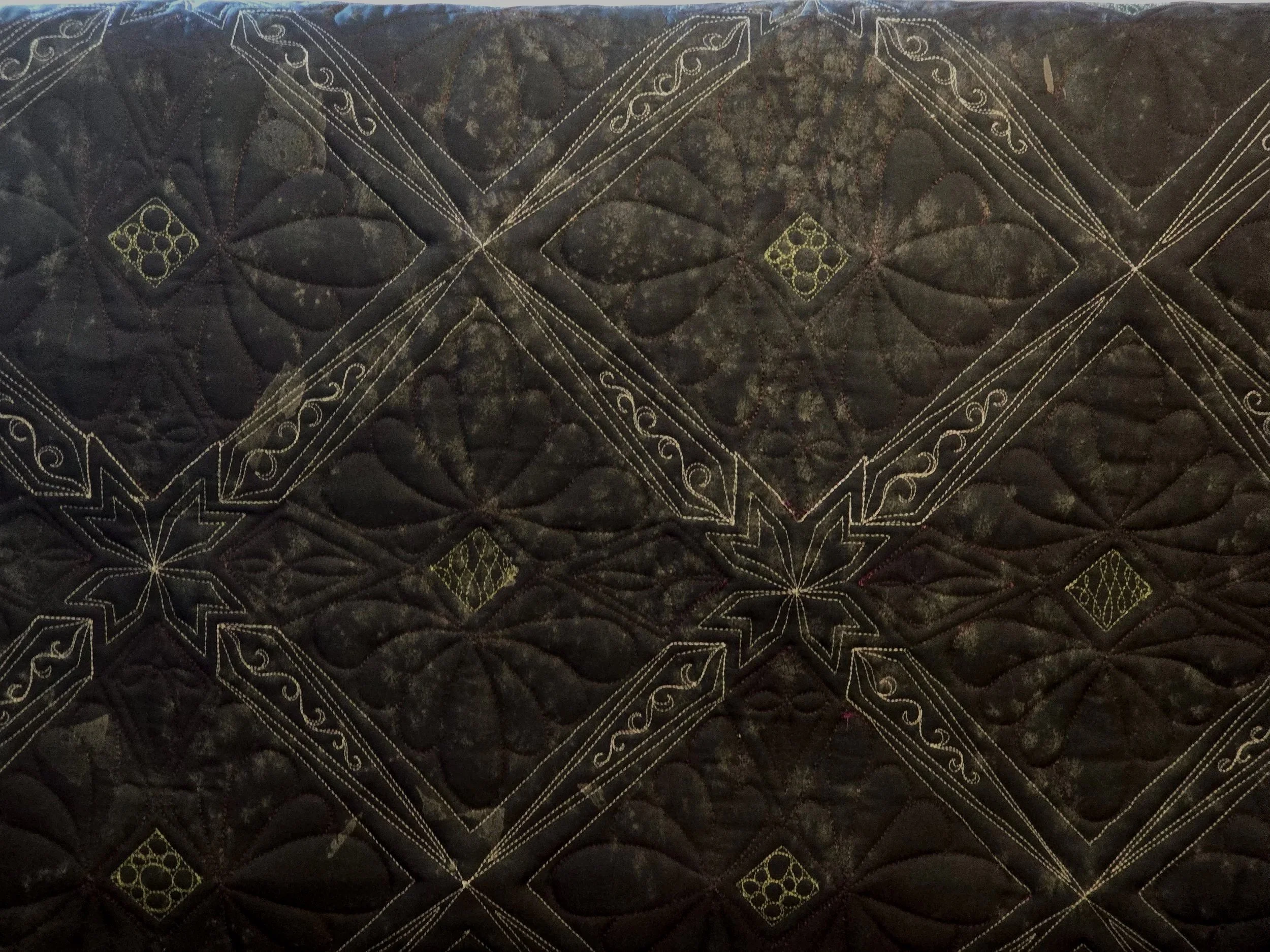 A dark-colored quilt with intricate stitched patterns and embroidered details, featuring floral and geometric motifs.