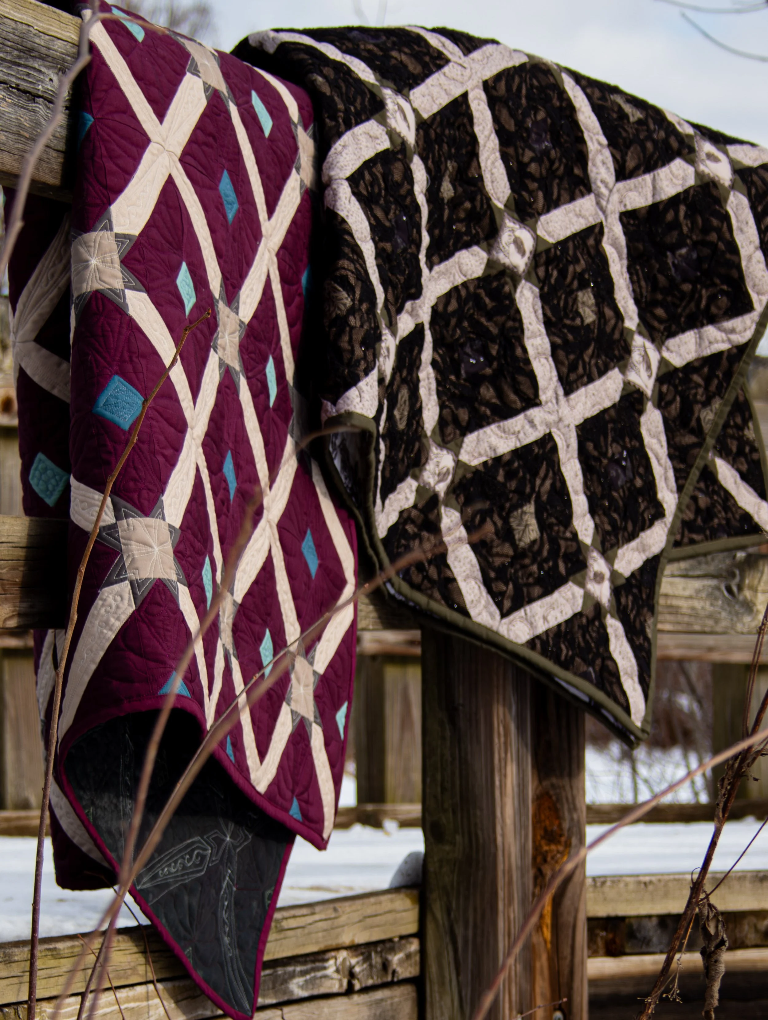 Two handmade quilts hanging on a wooden fence outdoors during winter, with snow on the ground.