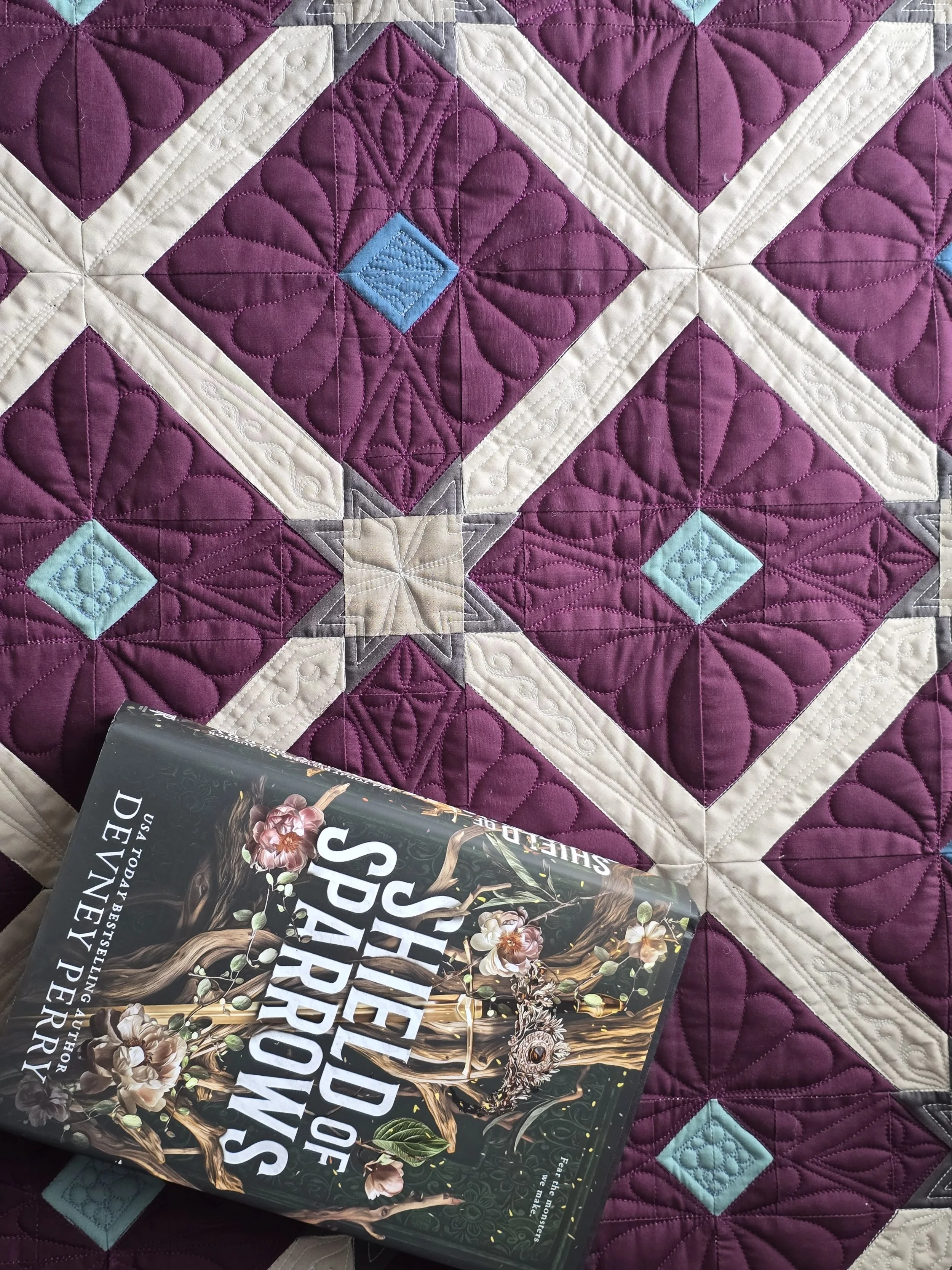 A quilt with deep purple, beige, brown/gray, and light blue geometric patterns and intricate free motion quilting stitching, and with a book titled 'Shield of Sparrows by Devney Perry", on it