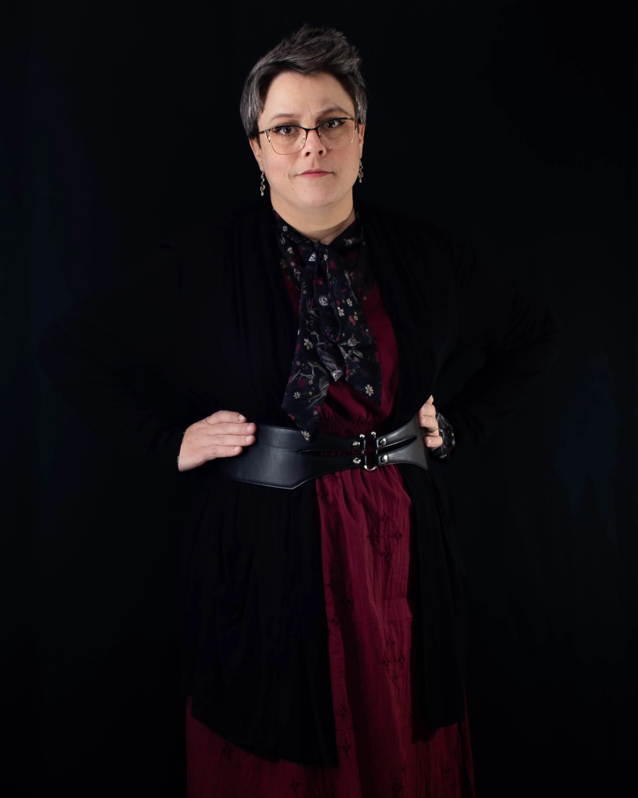 A woman with short gray hair and glasses posing with hands on hips against a black background, wearing a black jacket, a dark floral scarf, and a red dress with a wide black belt.