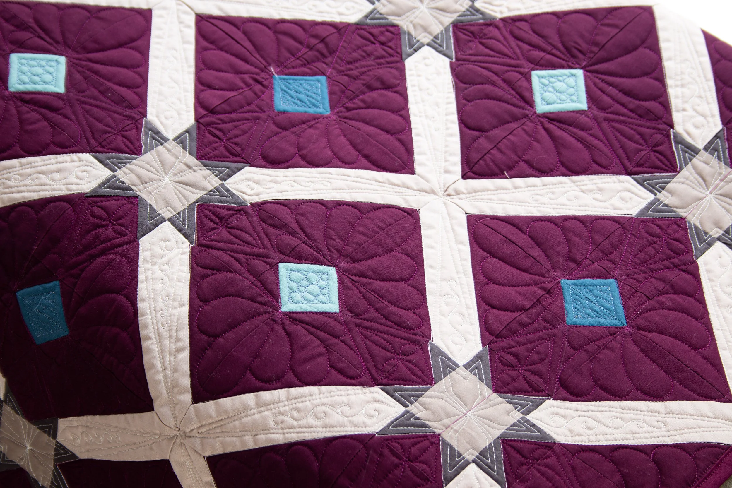 A close-up of a quilt with purple, white, gray, and light blue geometric patterns and intricate stitching.