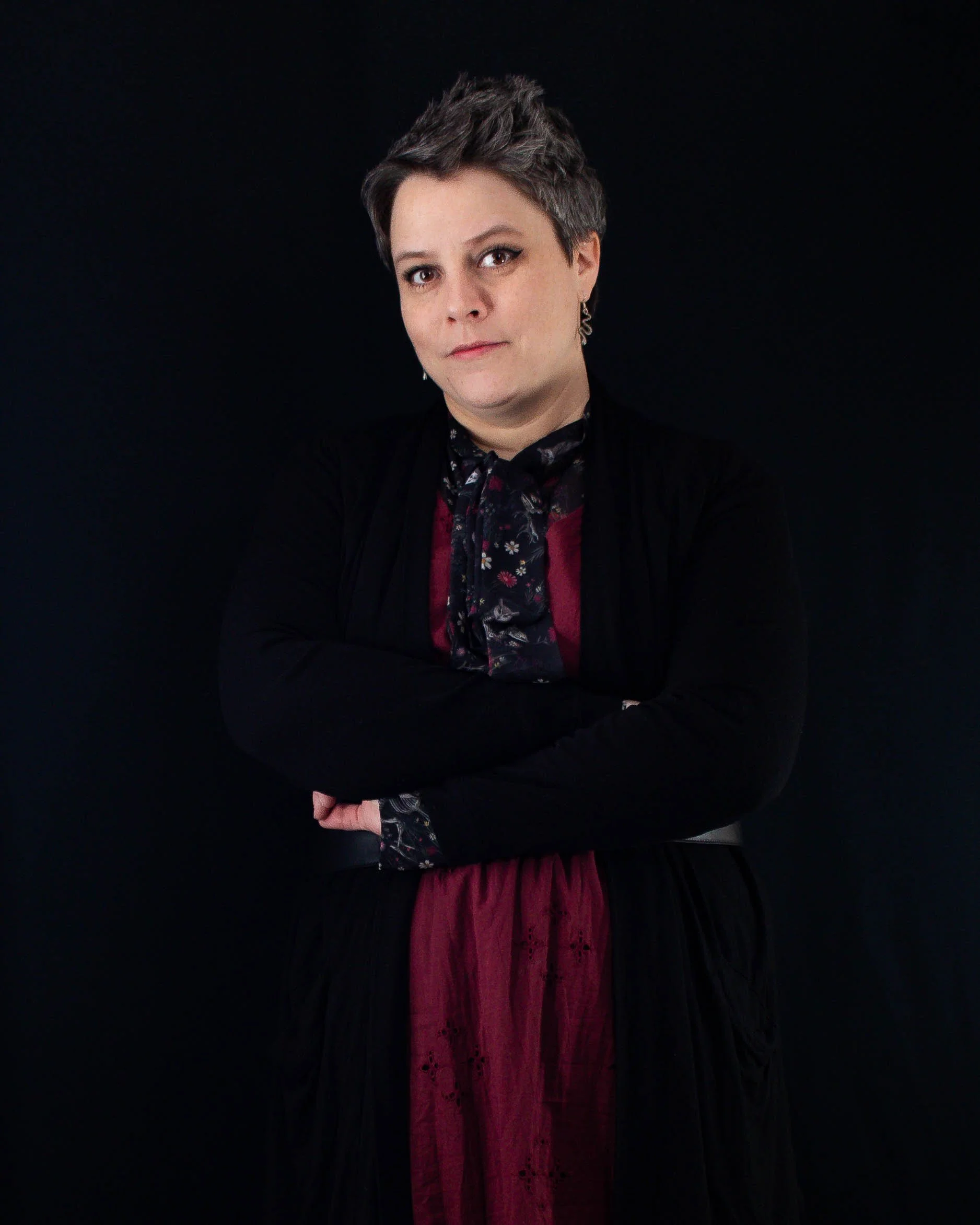 A woman with short, styled gray hair standing with arms crossed against a black background, wearing a black jacket and a maroon dress with a floral-patterned scarf.