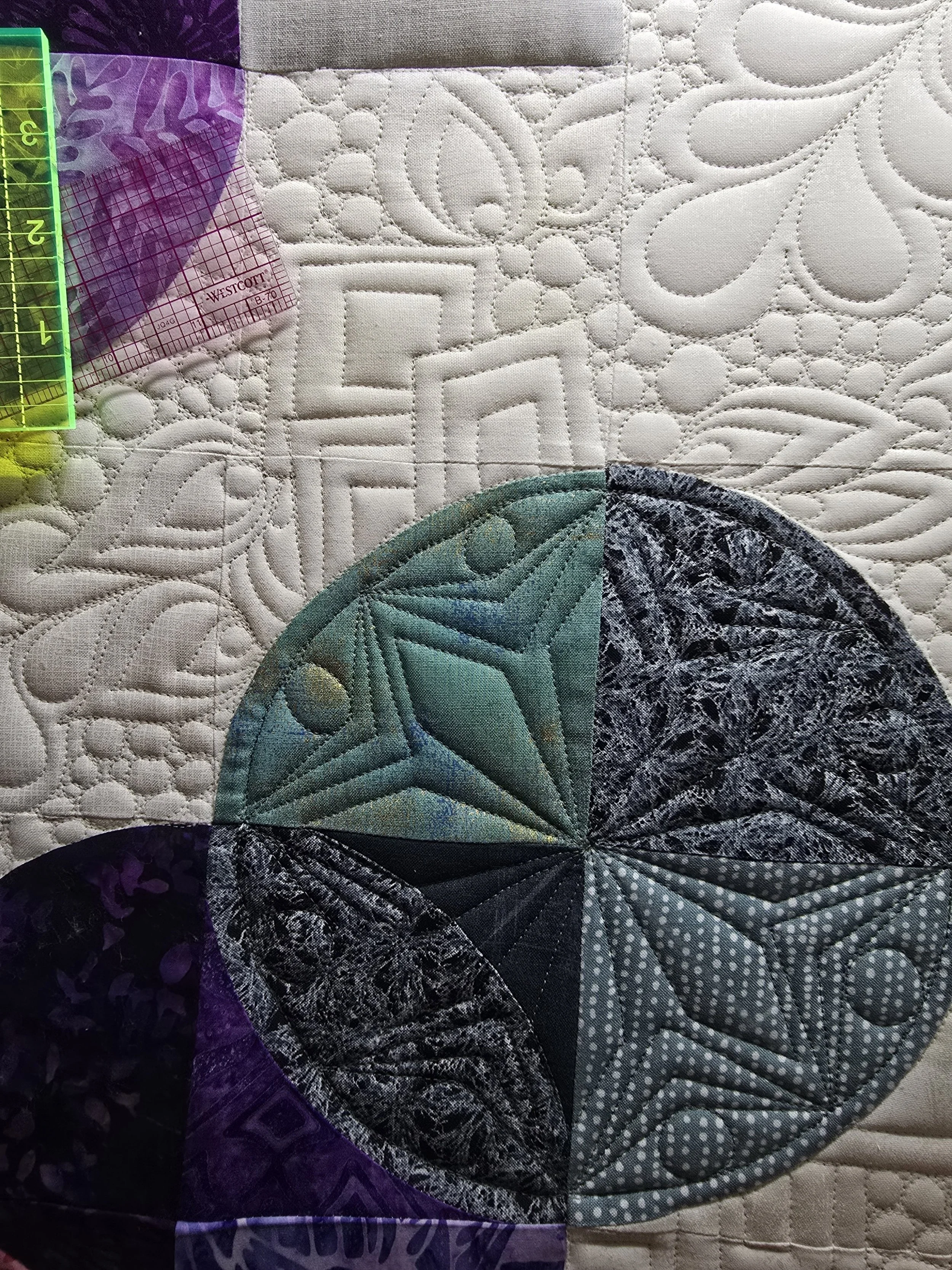Close-up of a quilt with intricate stitched patterns and a colorful circular fabric piece consisting of different fabric sections in green, black, and gray tones.
