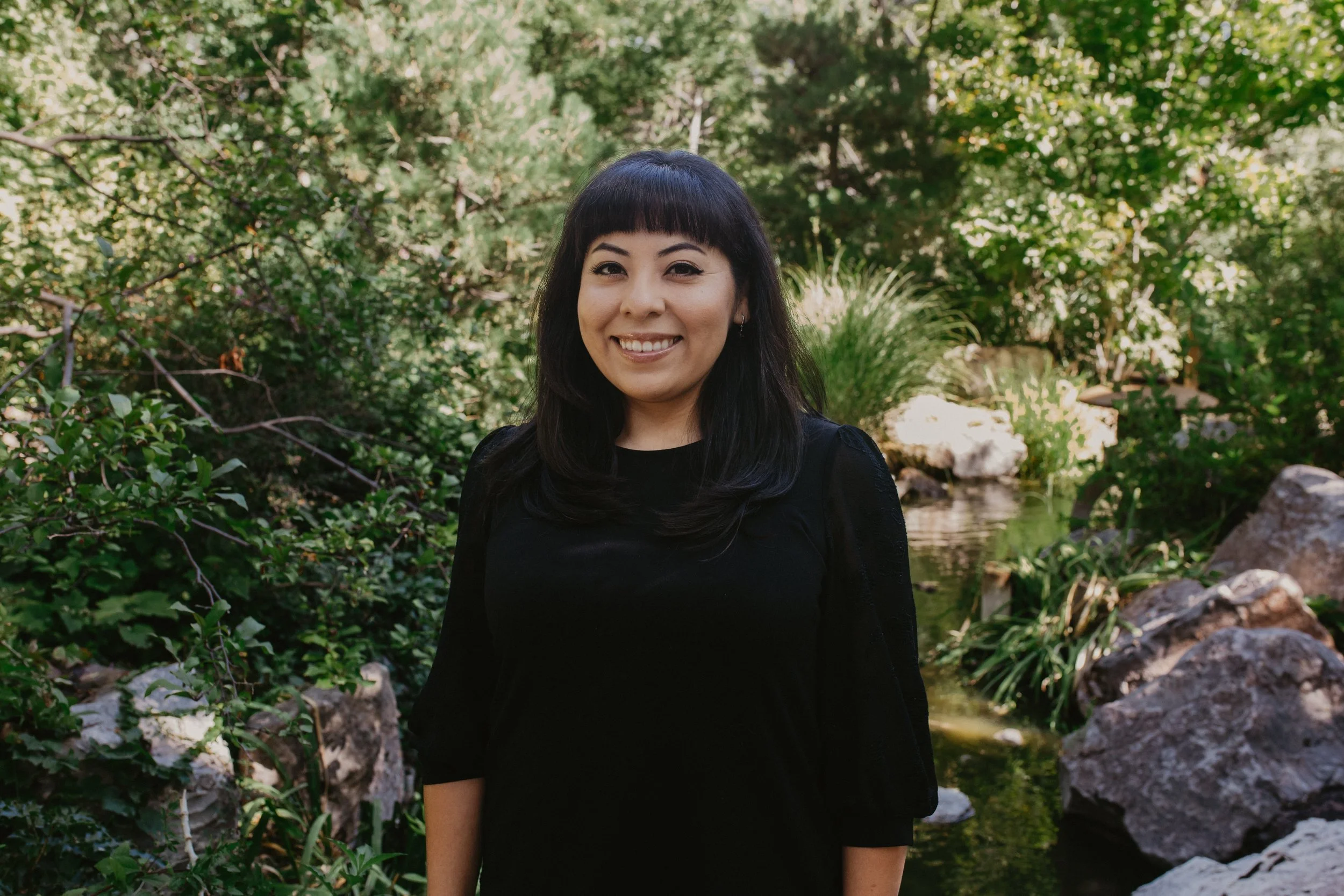 A woman with black hair and bangs smiling and standing outdoors near a pond surrounded by greenery and rocks.