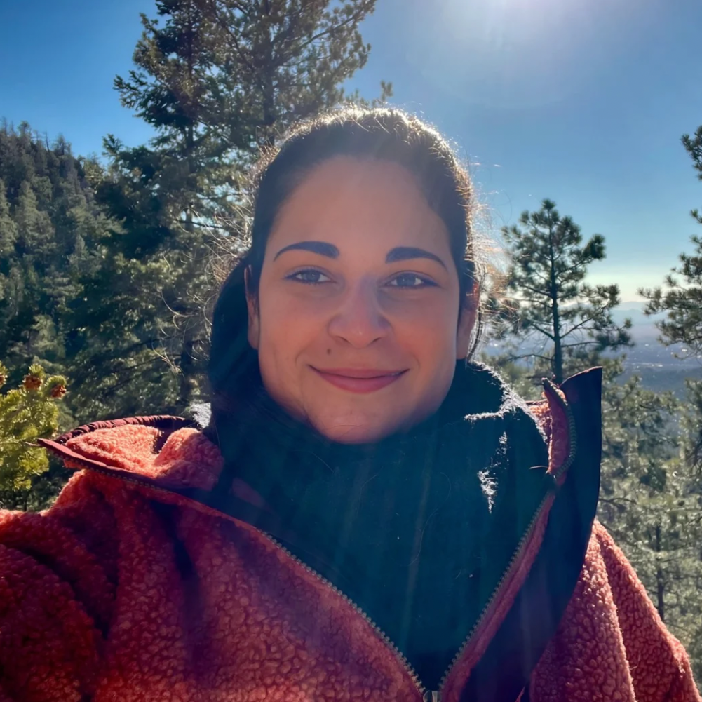 A woman smiling outdoors in a forested area with pine trees and mountains in the background, wearing a red fleece jacket and a black scarf, on a sunny day.