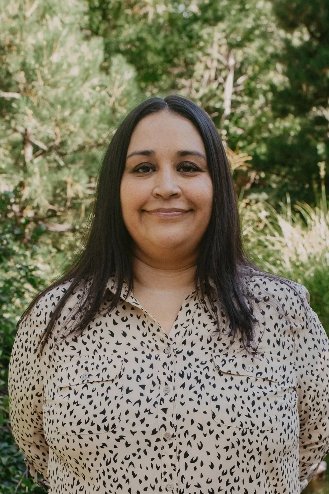 A woman with long black hair smiling outdoors in front of green trees, wearing a beige shirt with black abstract spots.