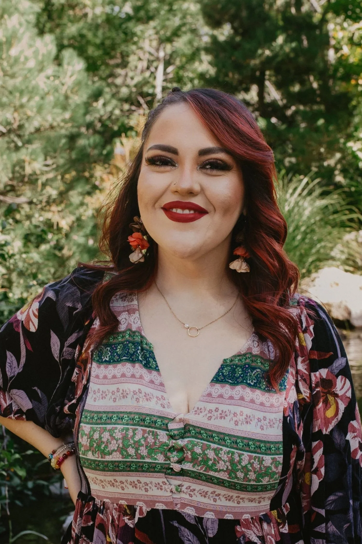 A woman outdoors with dark red hair, wearing floral earrings, a necklace, a patterned blouse with green, pink, and blue details, and a black floral jacket, smiling at the camera.