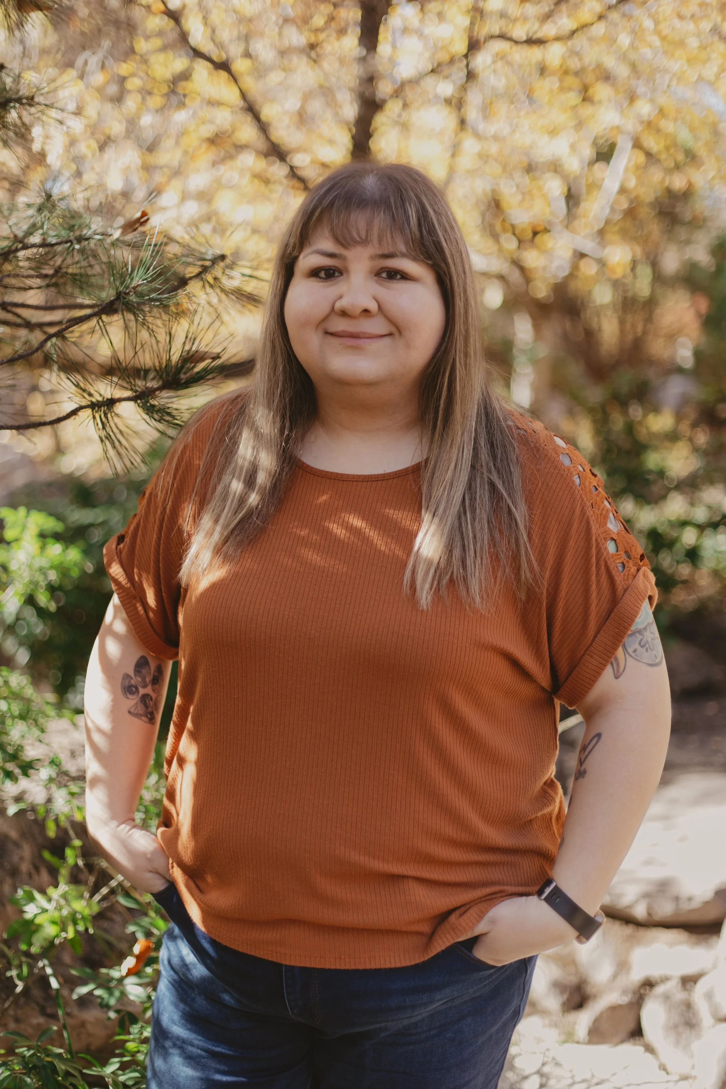 A woman standing outdoors in a forested area during fall, smiling and looking at the camera, wearing an orange short-sleeved top with cut-out details on the shoulders and tattoos on her arms.
