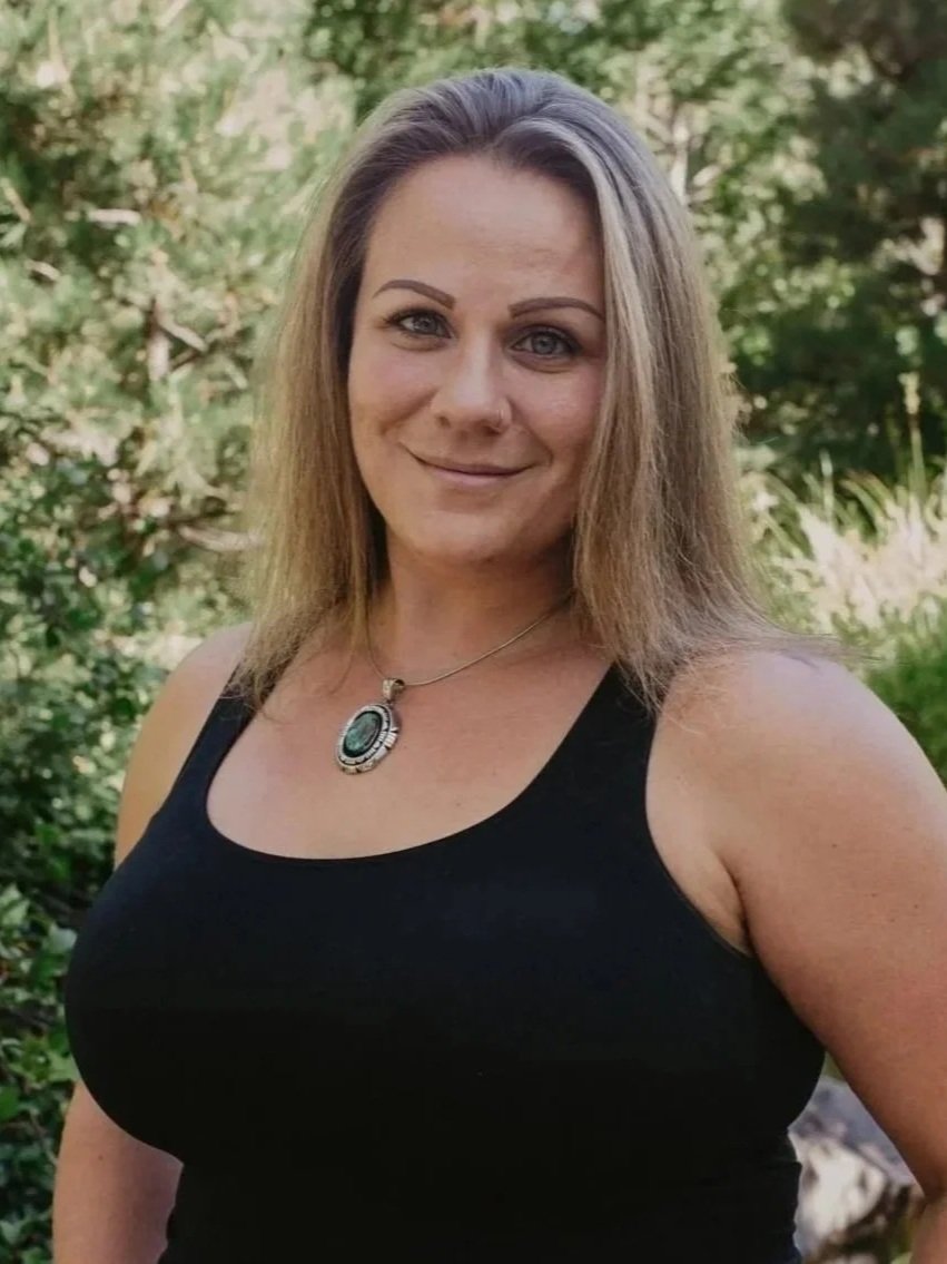 A woman with shoulder-length light brown hair, wearing a black sleeveless top and a silver necklace with an oval pendant, smiling outdoors with trees and greenery in the background.