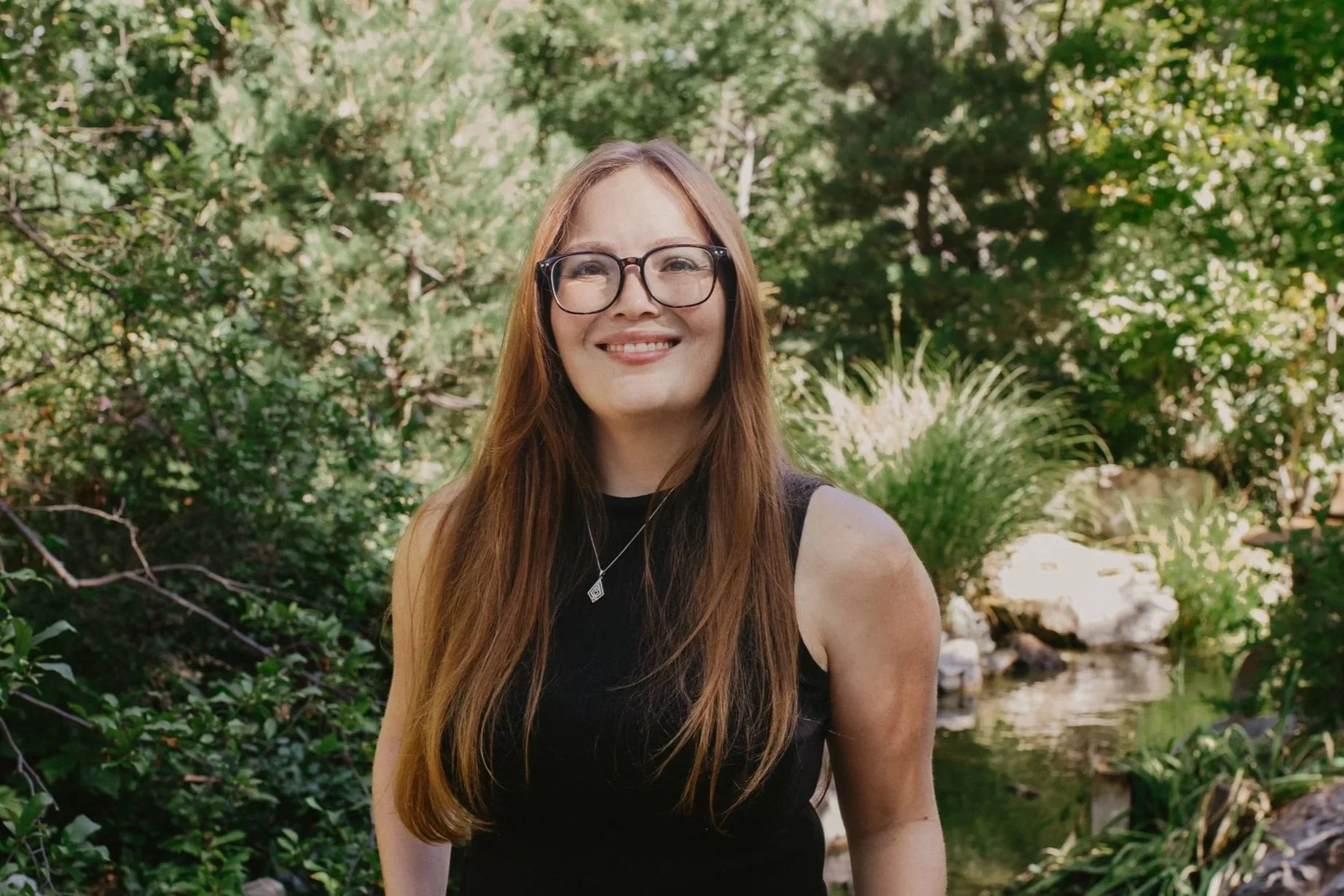 A woman with long red hair, wearing glasses, a black sleeveless top, and a necklace, smiling outdoors in a lush green garden with a stream in the background.