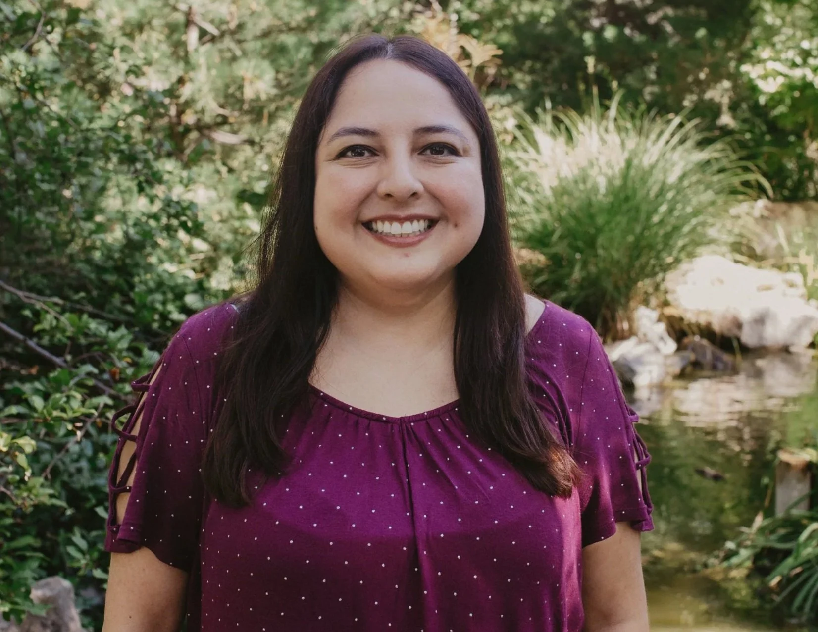 A woman with long dark hair, smiling, standing outdoors in a natural setting with greenery and a small pond in the background.