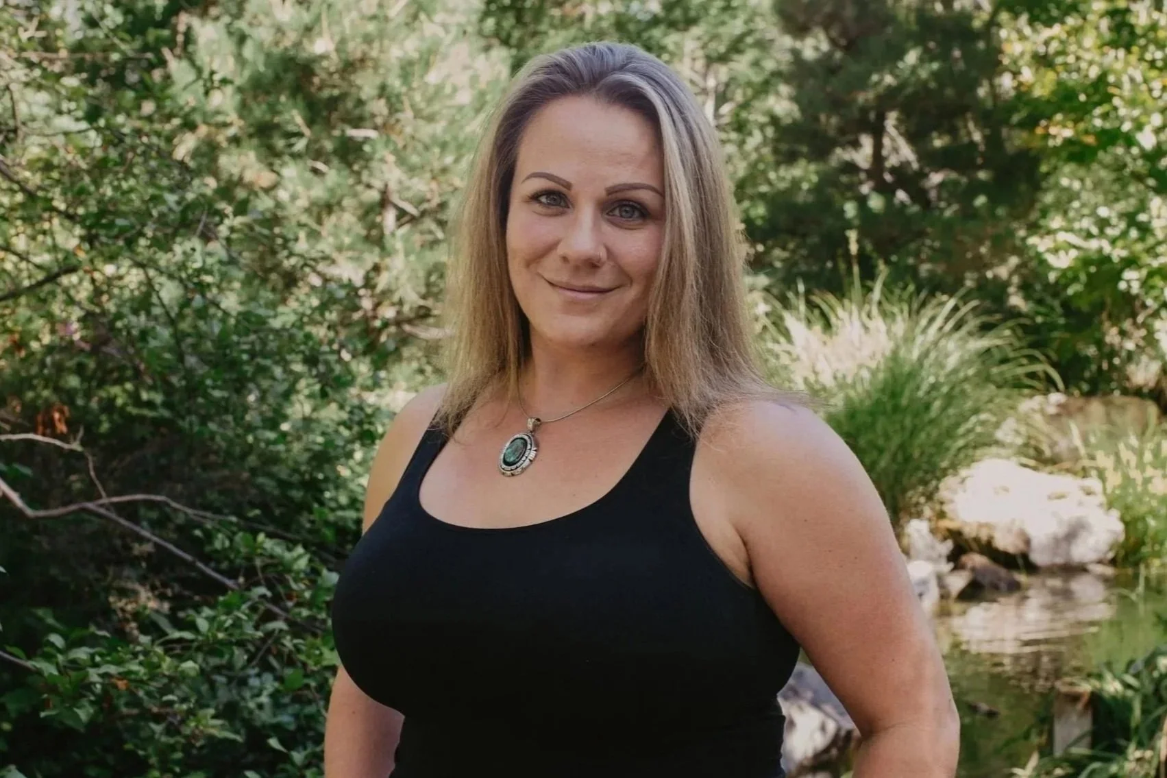 A woman in a black tank top posing outdoors in front of greenery and a small stream