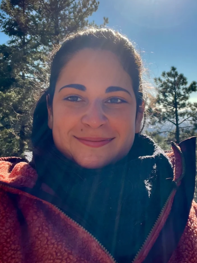 A woman with dark hair and eyebrows smiling outdoors with trees and a blue sky in the background.