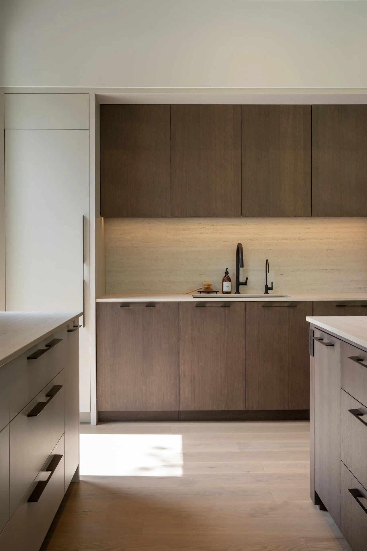 Cuisine moderne avec des armoires en bois clair et foncé, évier noir, robinetterie noire, comptoir en pierre beige, accessoires de vaisselle et savon sur le plan de travail.