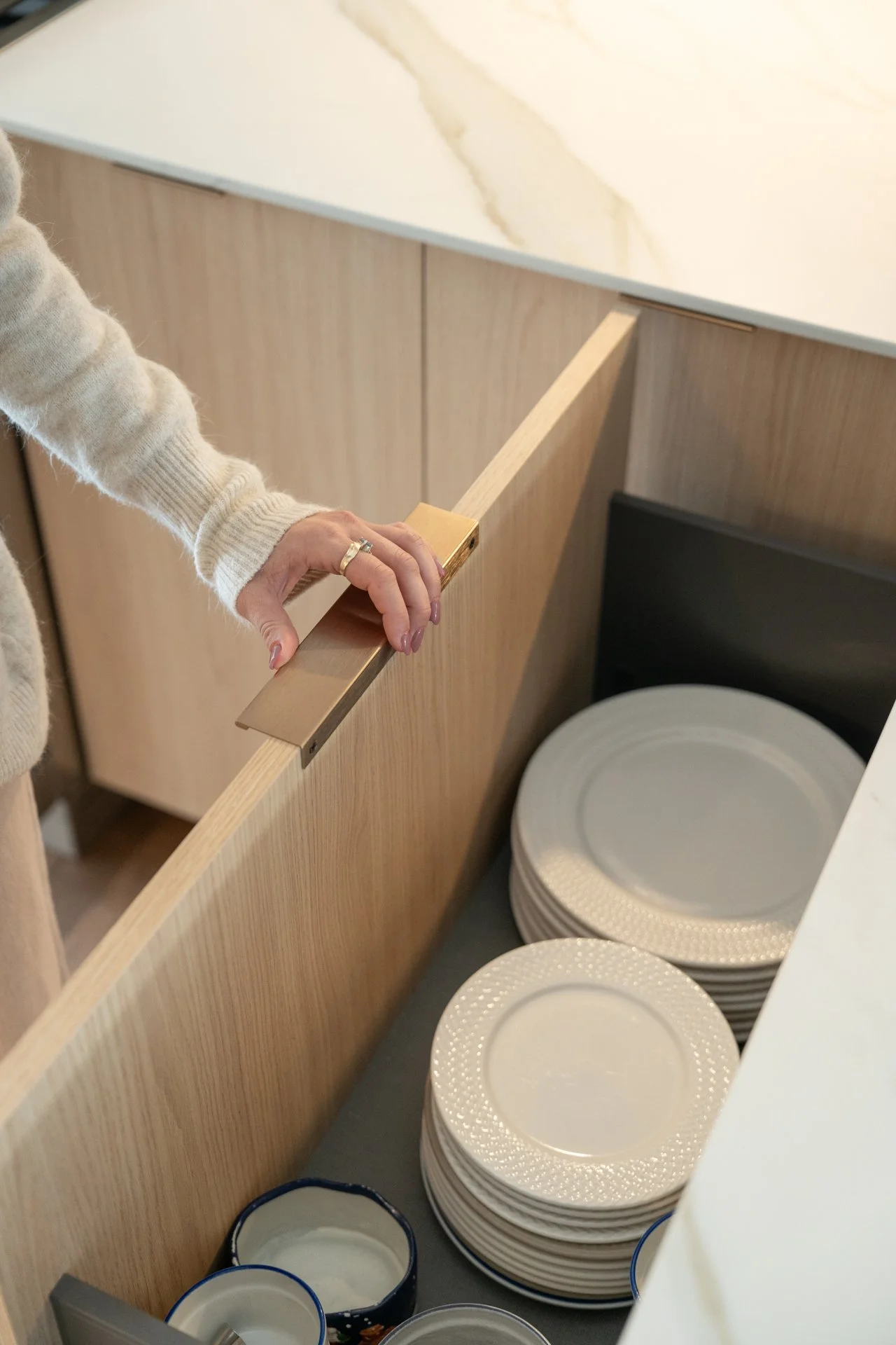 Une personne dépose une assiette dans une cuisine moderne avec un comptoir en marbre, des placards en bois clair et des piles d'assiettes blanches à motif. Il y a aussi des bols en porcelaine blanche avec un contour bleu.