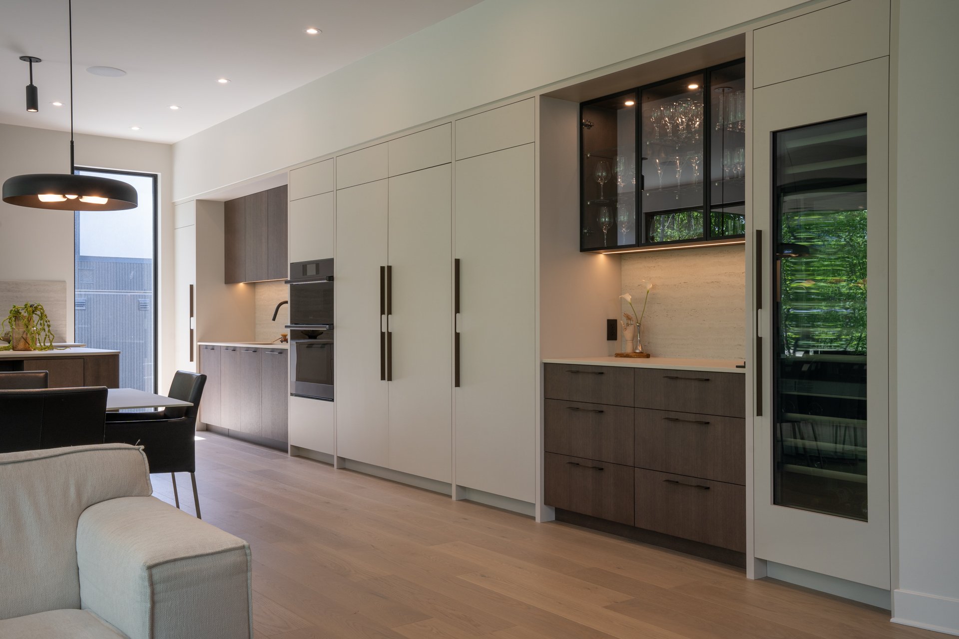 Intérieur moderne d'une cuisine avec des armoires blanches et en bois, une grande fenêtre laissant entrer de la lumière naturelle, et un espace repas avec des chaises noires.