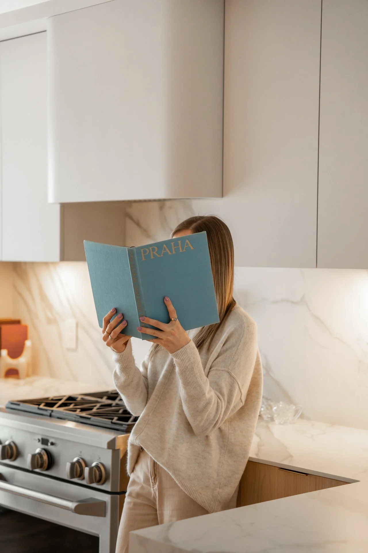 Une femme tient un livre intitulé "PRAHA" dans une cuisine moderne, debout près d'une cuisinière.