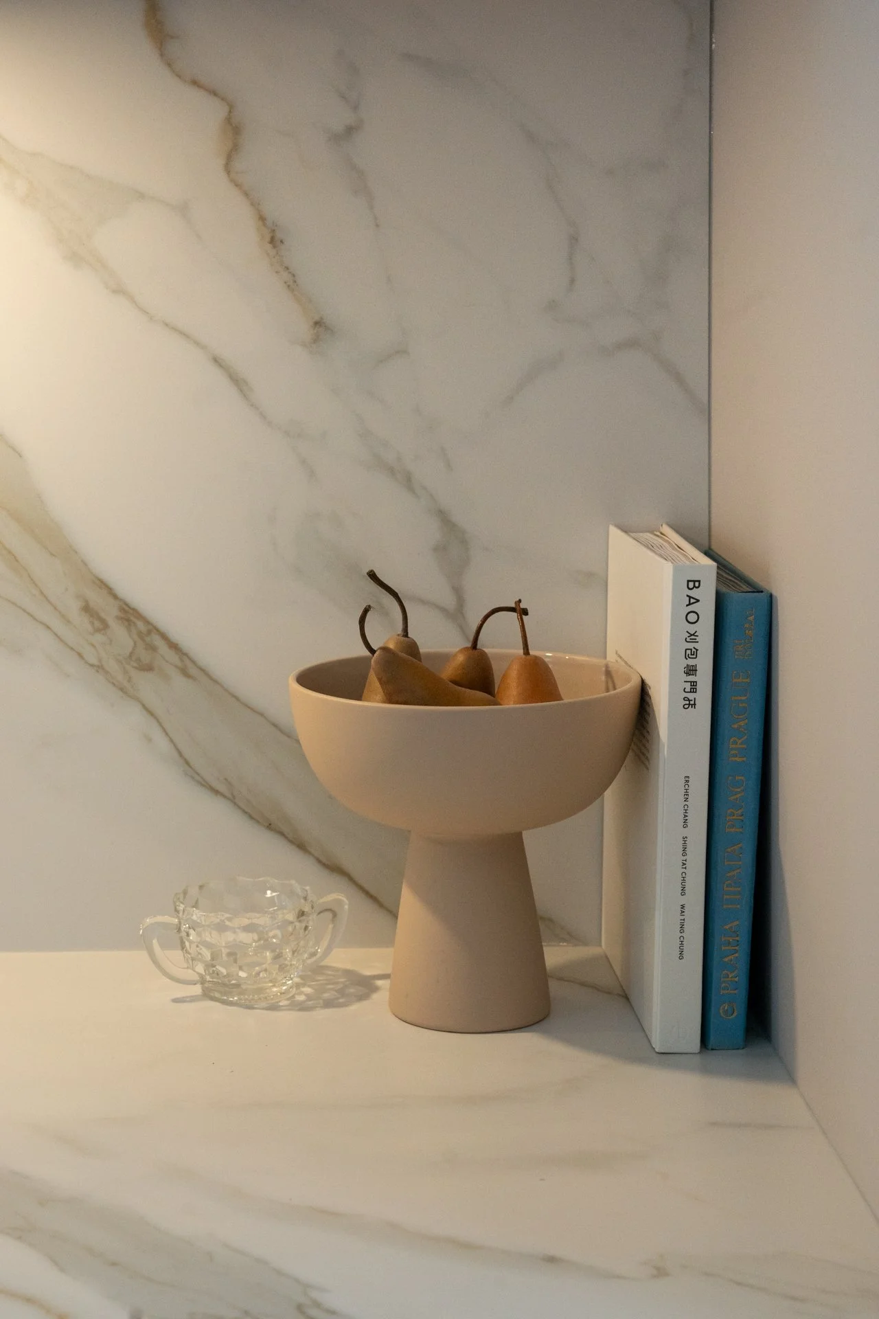 Une table en marbre blanc avec une coupe de fruits en céramique beige contenant des poires, un livre blanc et deux livres bleus empilés à droite. Une tasse en verre à motif est placée devant la coupe de fruits.