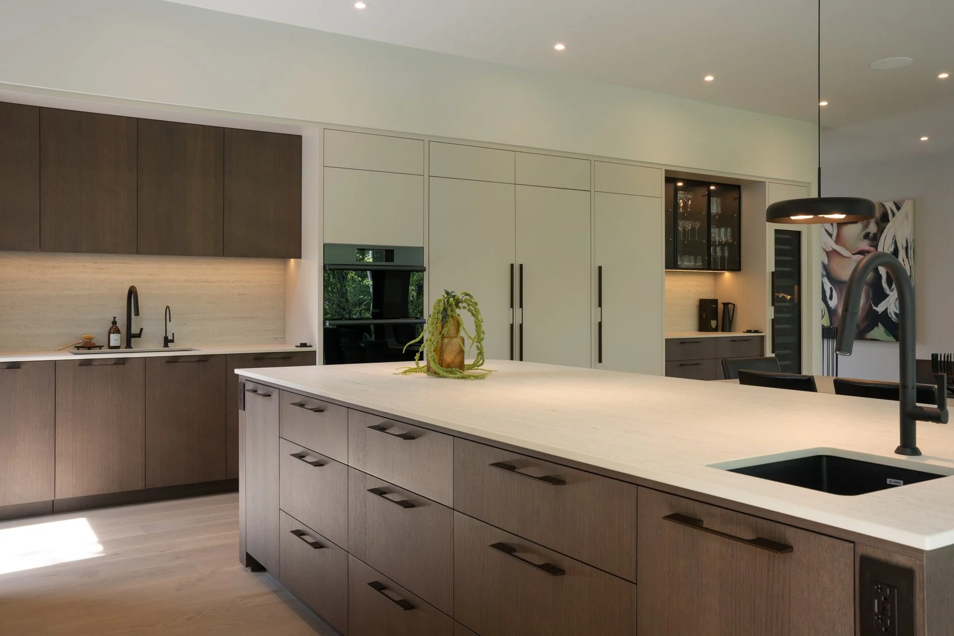 Cuisine moderne avec îlot en bois clair, comptoir en pierre blanche, et appareils intégrés. Mur en peinture marron foncé et blanc avec éclairage encastré. Décoration minimaliste avec une plante sur l'îlot.