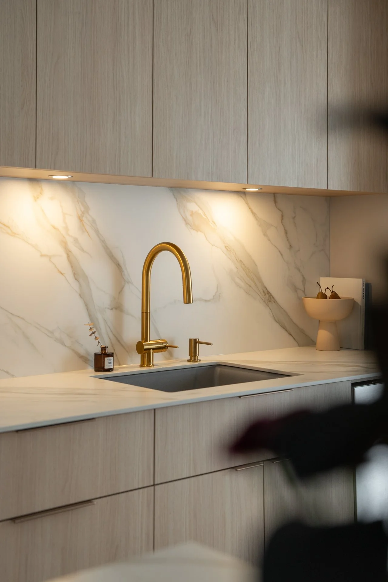 Cuisine moderne avec un plan de travail en marbre blanc, un évier en acier inoxydable, un robinet doré, des armoires en bois clair, un mur en marbre avec des veines dorées, et un décor minimaliste avec un bol blanc contenant des poires.