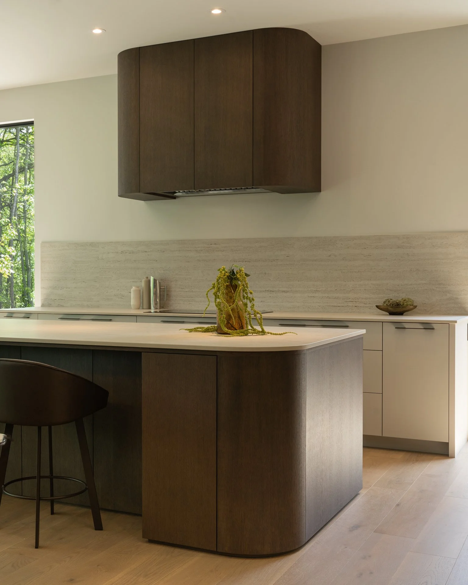 Cuisine moderne avec îlot en bois foncé, plan de travail en marbre clair, meubles blancs, fenêtre avec vue sur des arbres, décoration simple avec vase et livres.