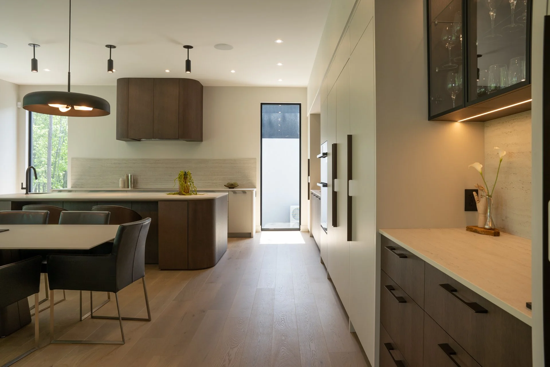 Cuisine moderne avec îlot central, meubles en blanc et bois, éclairage encastré, table avec chaises noires, porte vitrée laissant entrer la lumière, décor minimaliste avec vase et fleurs.