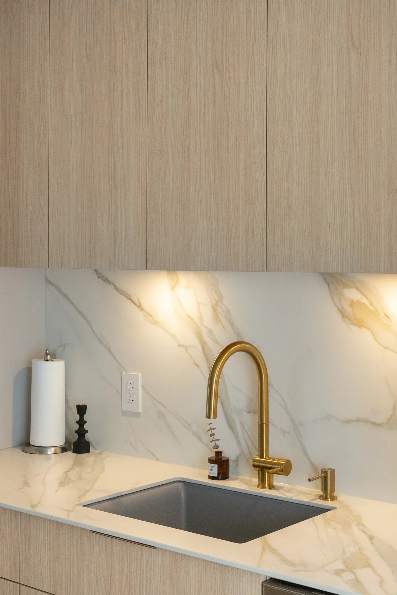 Cuisine moderne avec un comptoir en marbre beige avec veines dorées, une évier en acier inoxydable, un robinet doré, une lampe de papier blanc, une bouteille de savon, et un mur en marbre avec des meubles en bois clair au-dessus.