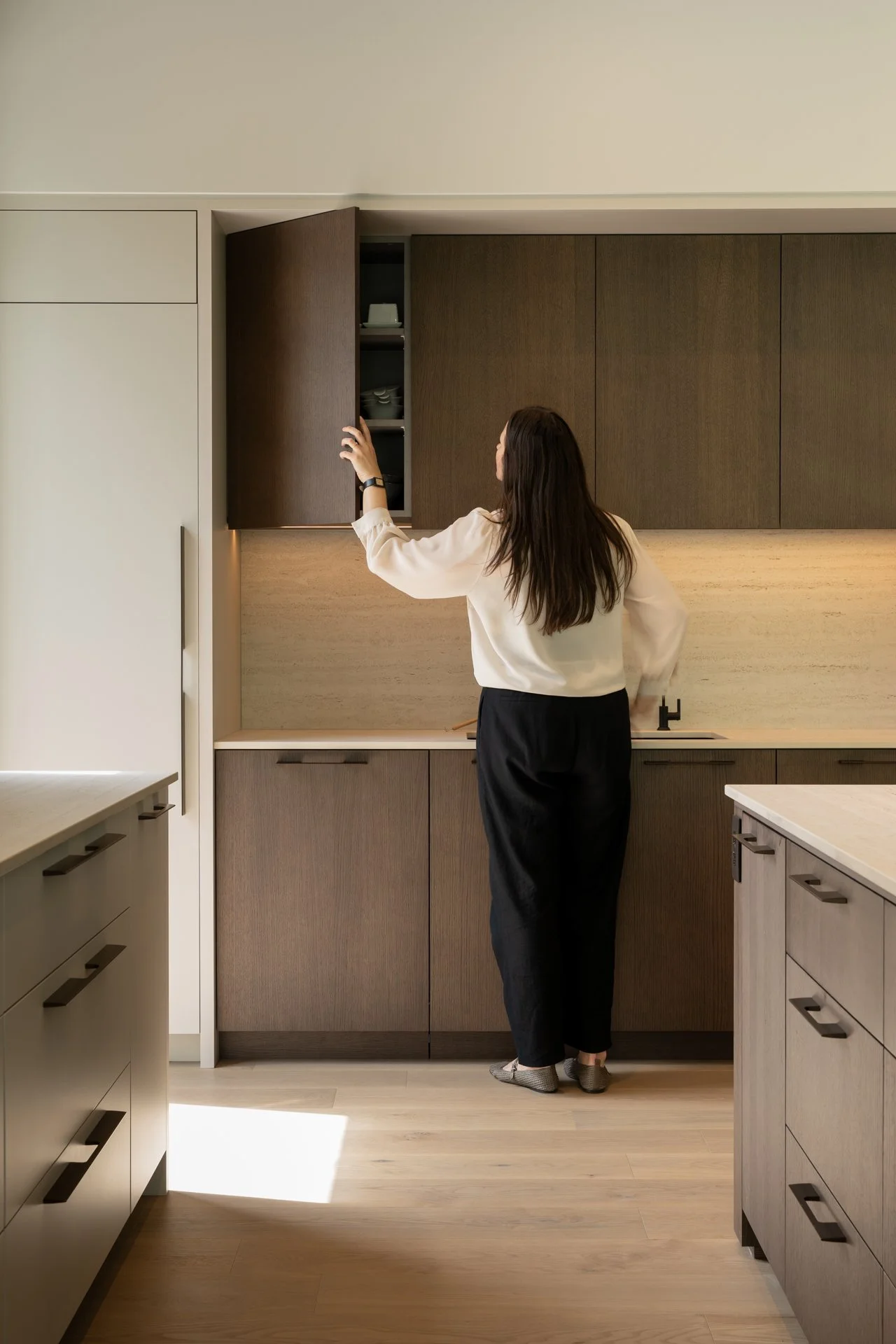 Femme en vêtements décontractés ouvrant un placard de cuisine moderne en bois foncé, dans une cuisine minimaliste bien éclairée.