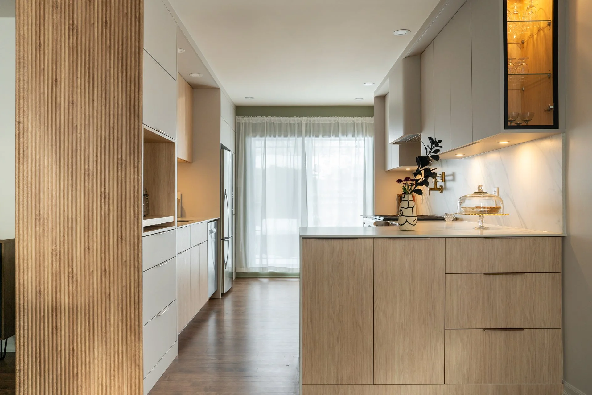 Cuisine moderne avec plan de travail en marbre, placards en bois clair, et étagère vitrée avec verre et vaisselle, éclairage intégré et grande fenêtre avec rideau blanc.