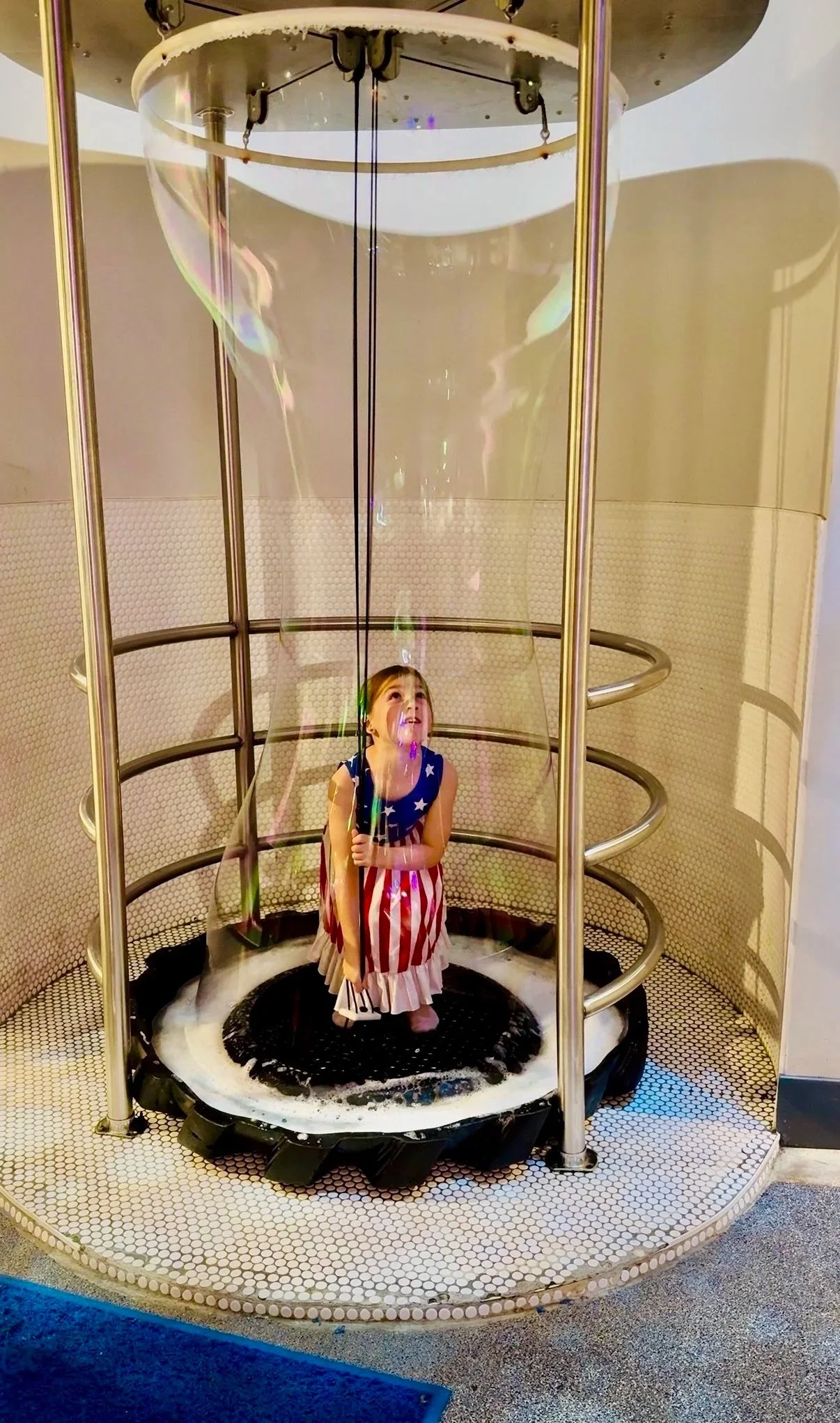 A young girl dressed in an American flag-themed dress standing on a small trampoline inside a bubble-making machine, looking up at the falling soap bubbles.
