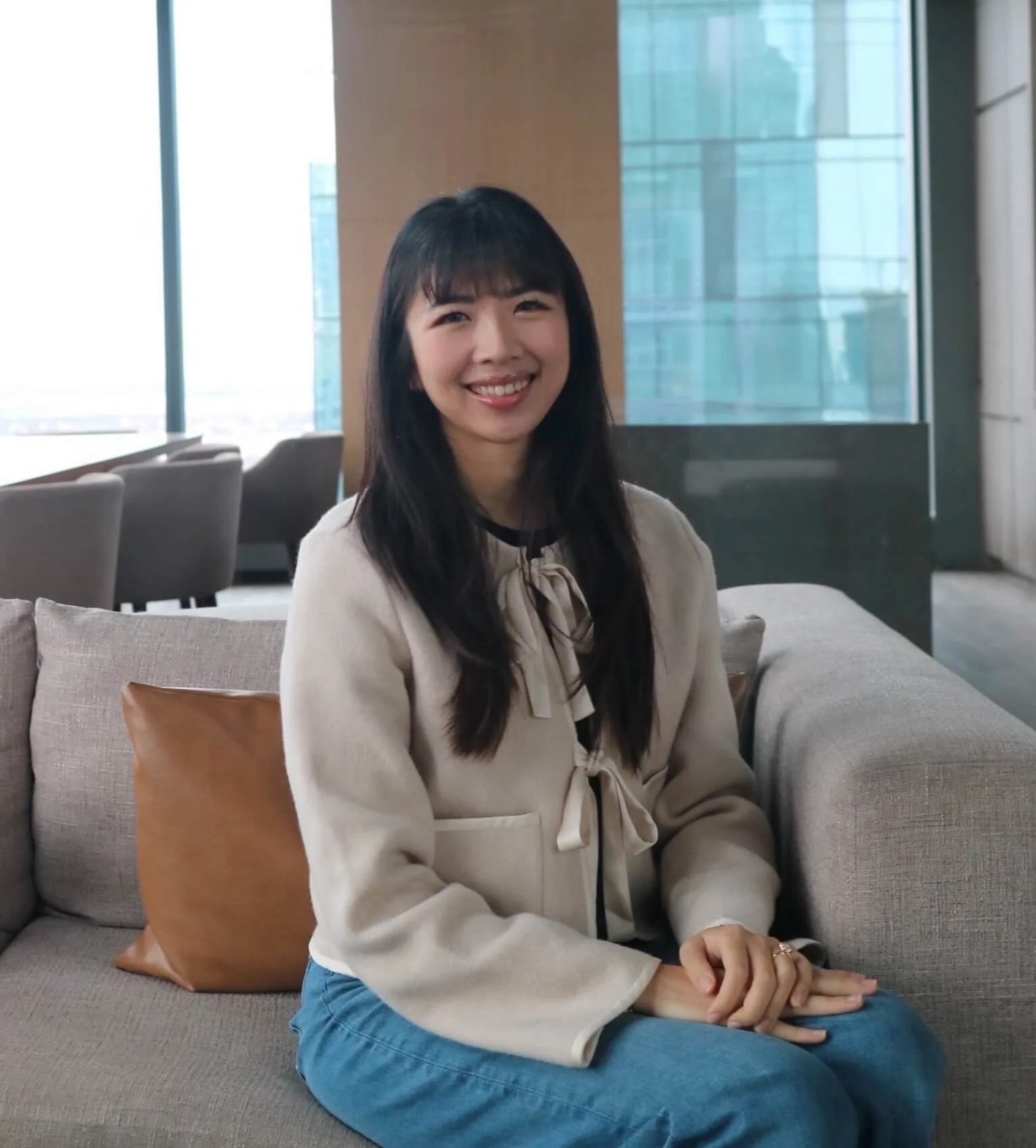 A young Asian woman with long dark hair and bangs, smiling, sitting on a gray couch with a brown pillow, in a modern office lobby with large windows and city views, wearing a cream-colored blazer with a bow tie detail and blue jeans.
