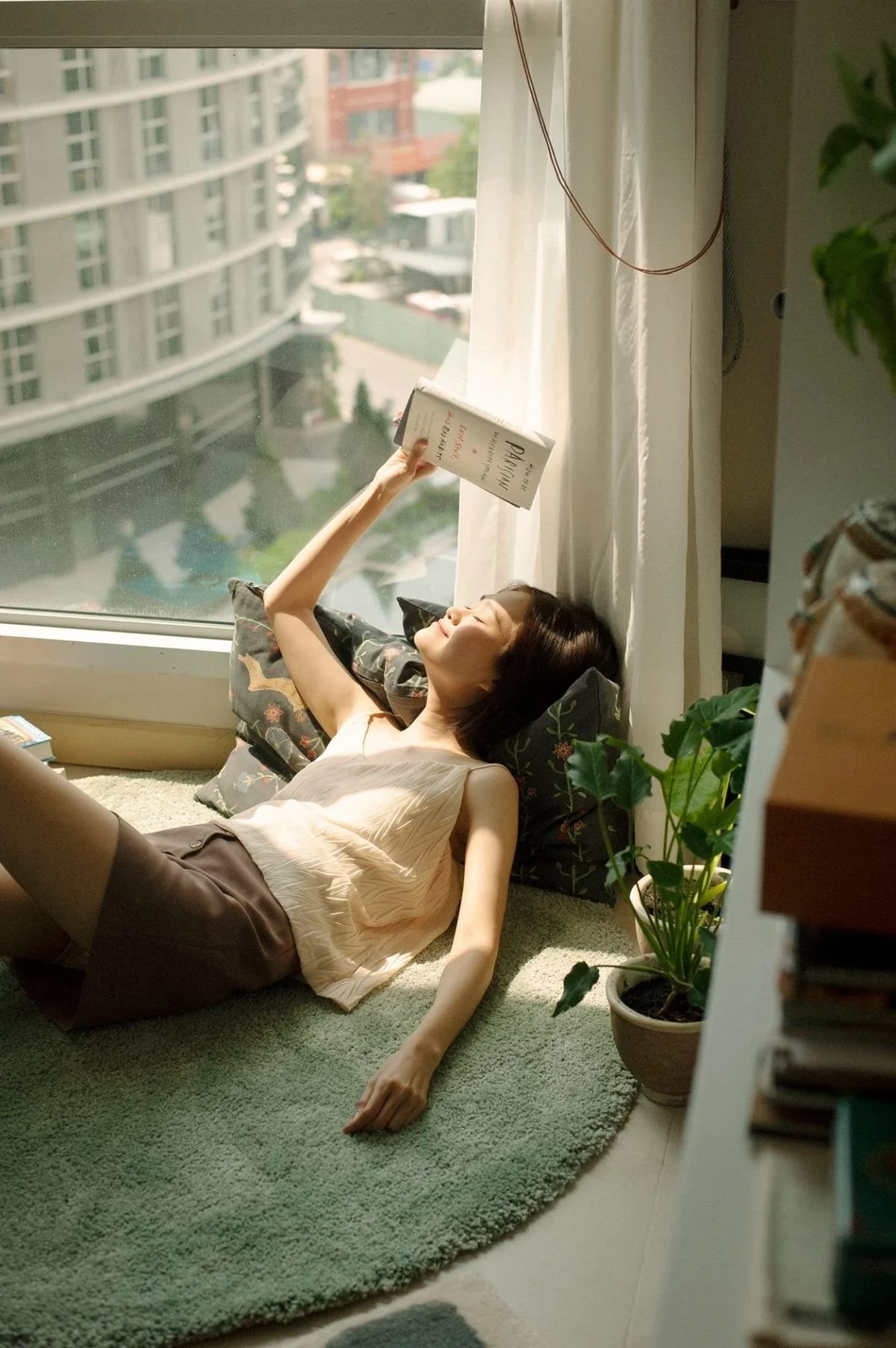 A woman is lying on a green rug by a window, reading a book, with sunlight shining on her face. She is surrounded by a pillow and a potted plant, with a view of buildings outside.