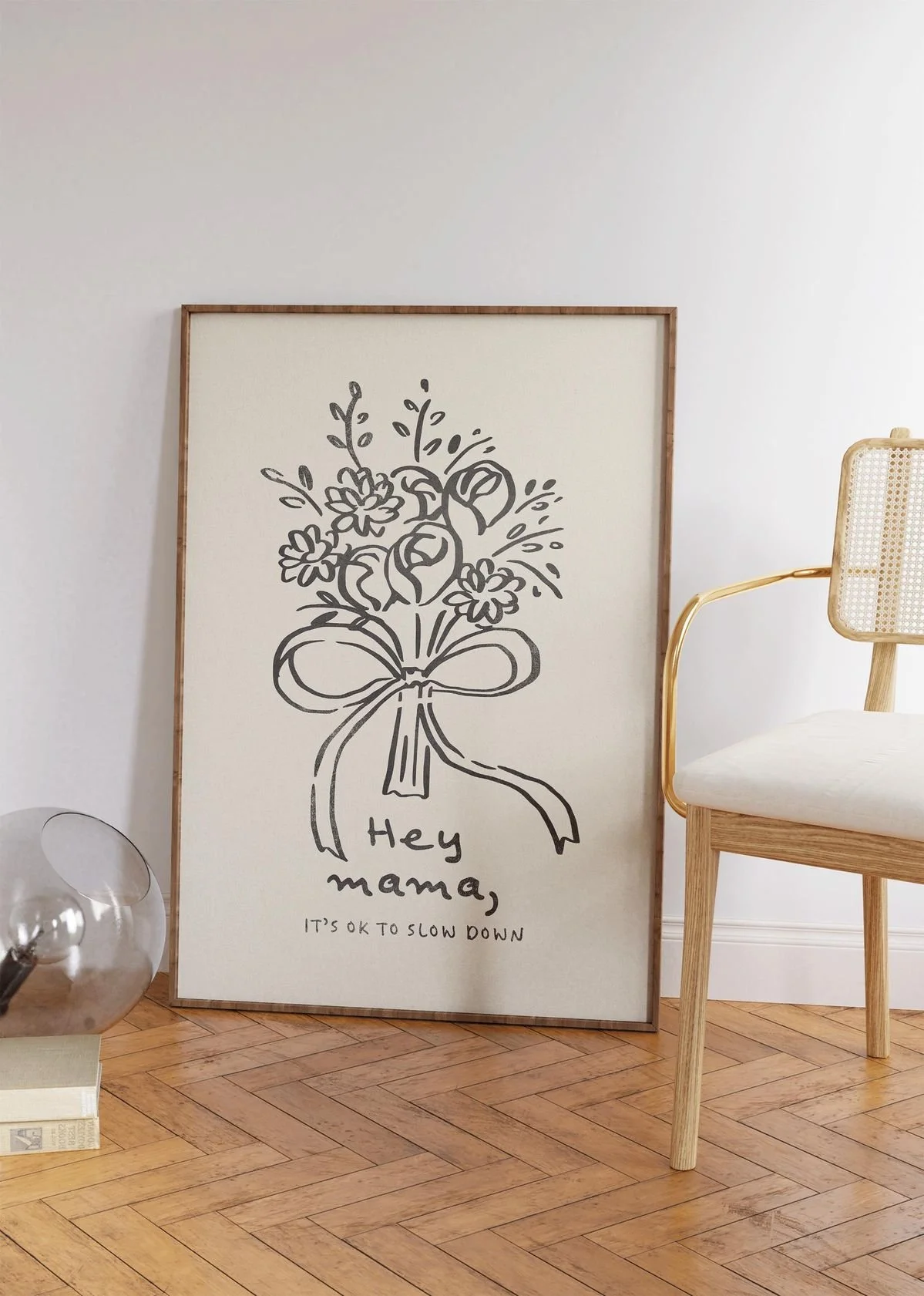 Framed wall art with a line drawing of a bouquet of flowers and a bow. Text reads, 'Hey mama, it's okay to slow down.' Beside the art, there is a wooden chair with a rattan backrest, and a glass orb decanter on a book on the floor.