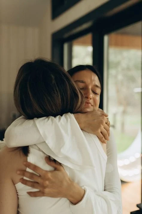 Two women hugging each other inside a house with large windows, one woman visibly emotional.