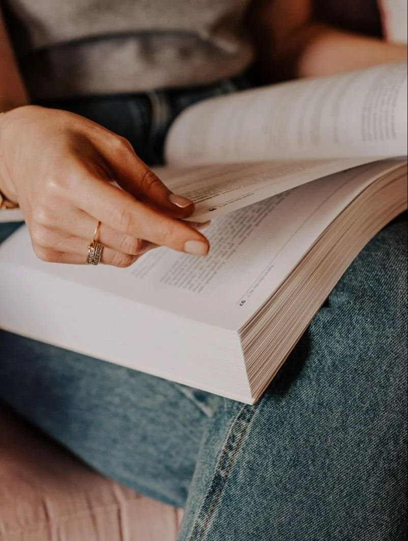 A person is sitting on a couch, reading a large, open book, with visible text on the pages, wearing a ring on their finger and jeans.
