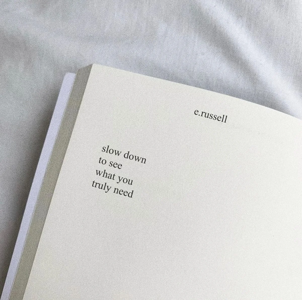 Close-up of an open white book with the title 'slow down to see what you truly need' by e. russell on a light grey surface.