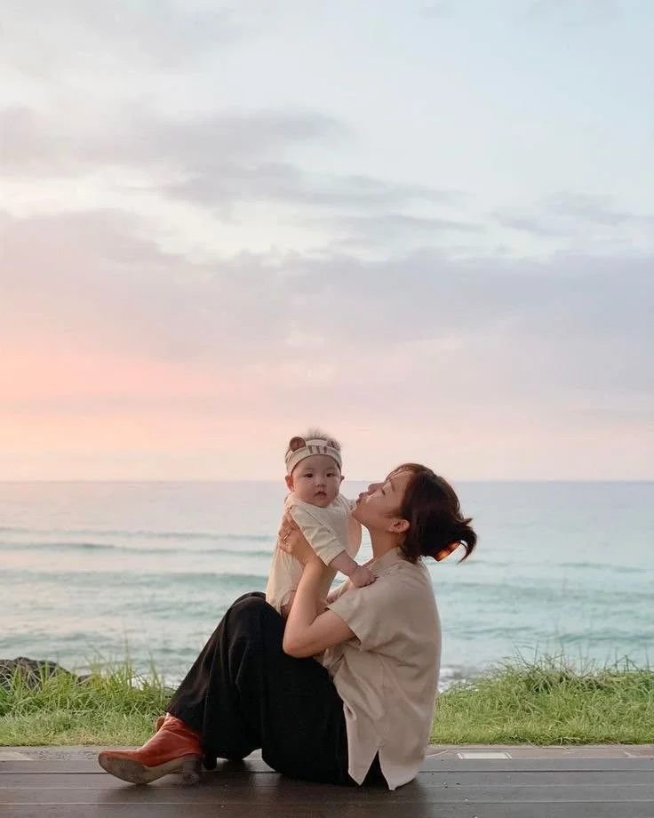 A woman holding a baby while sitting on a wooden deck near the beach during sunset, with the ocean and sky in the background.