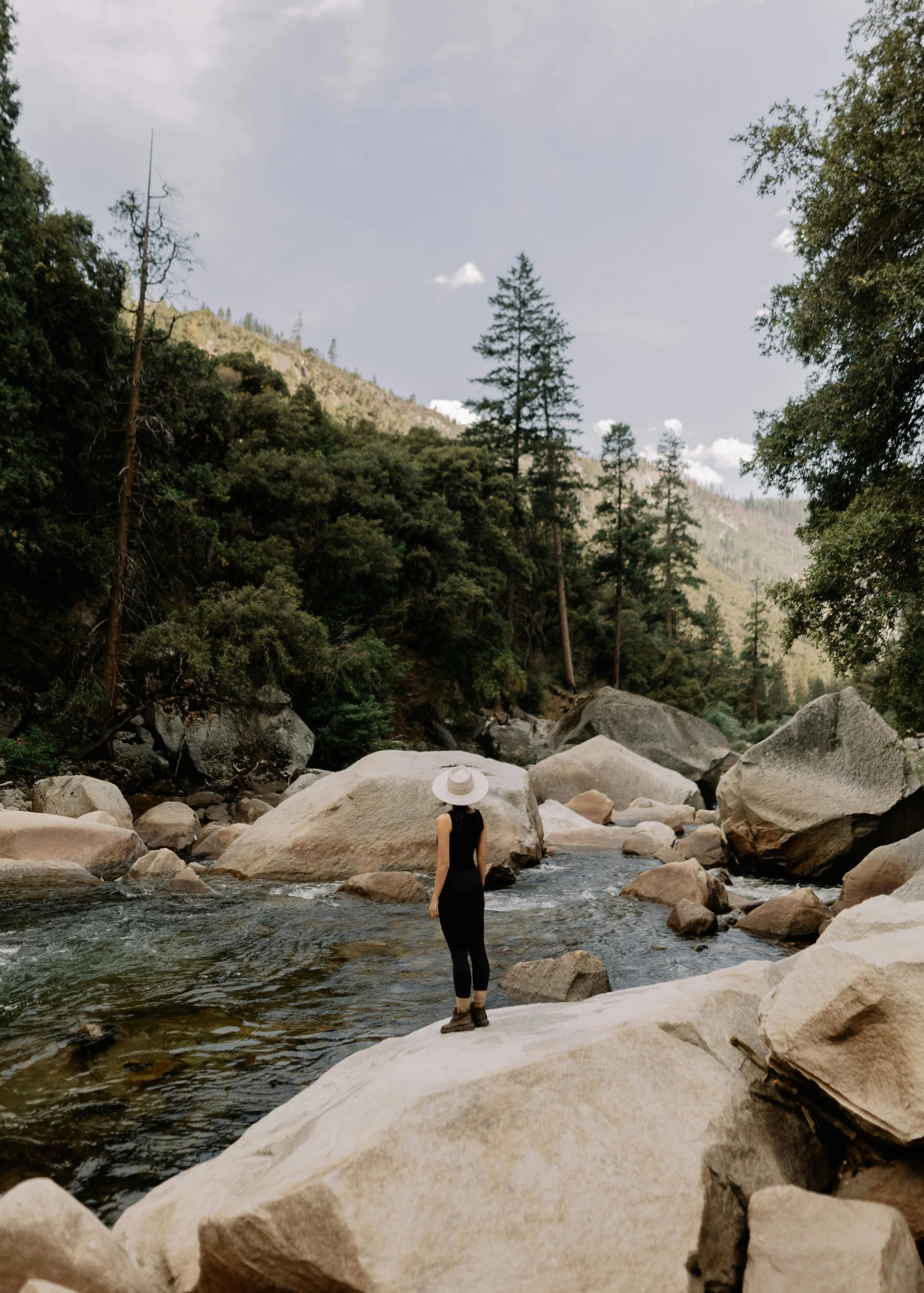 A woman in black clothing and hiking boots stands on a large rock in a shallow river, with tall trees and mountains in the background, under a partly cloudy sky.