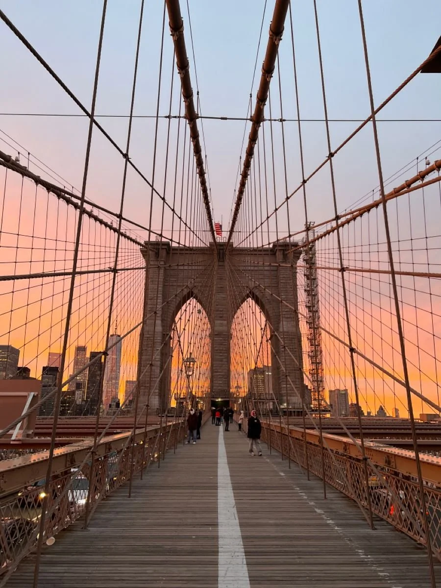View of Brooklyn Bridge at sunset with pedestrians walking on the bridge walkway, Brooklyn skyline in the background, and the bridge's cables and towers in focus.