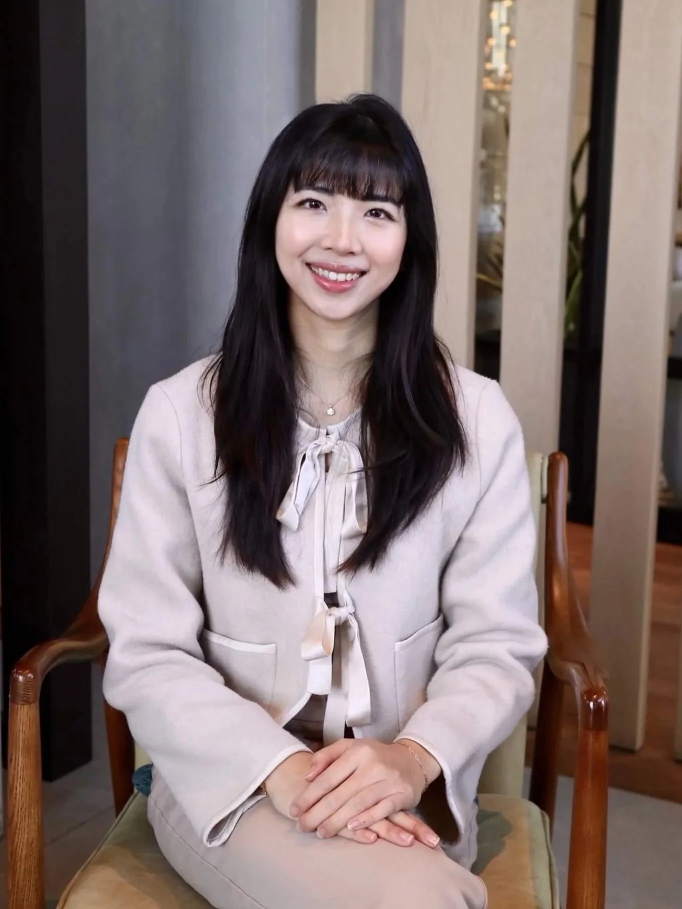 A woman with long dark hair and bangs, smiling, sitting in a chair in an indoor setting.