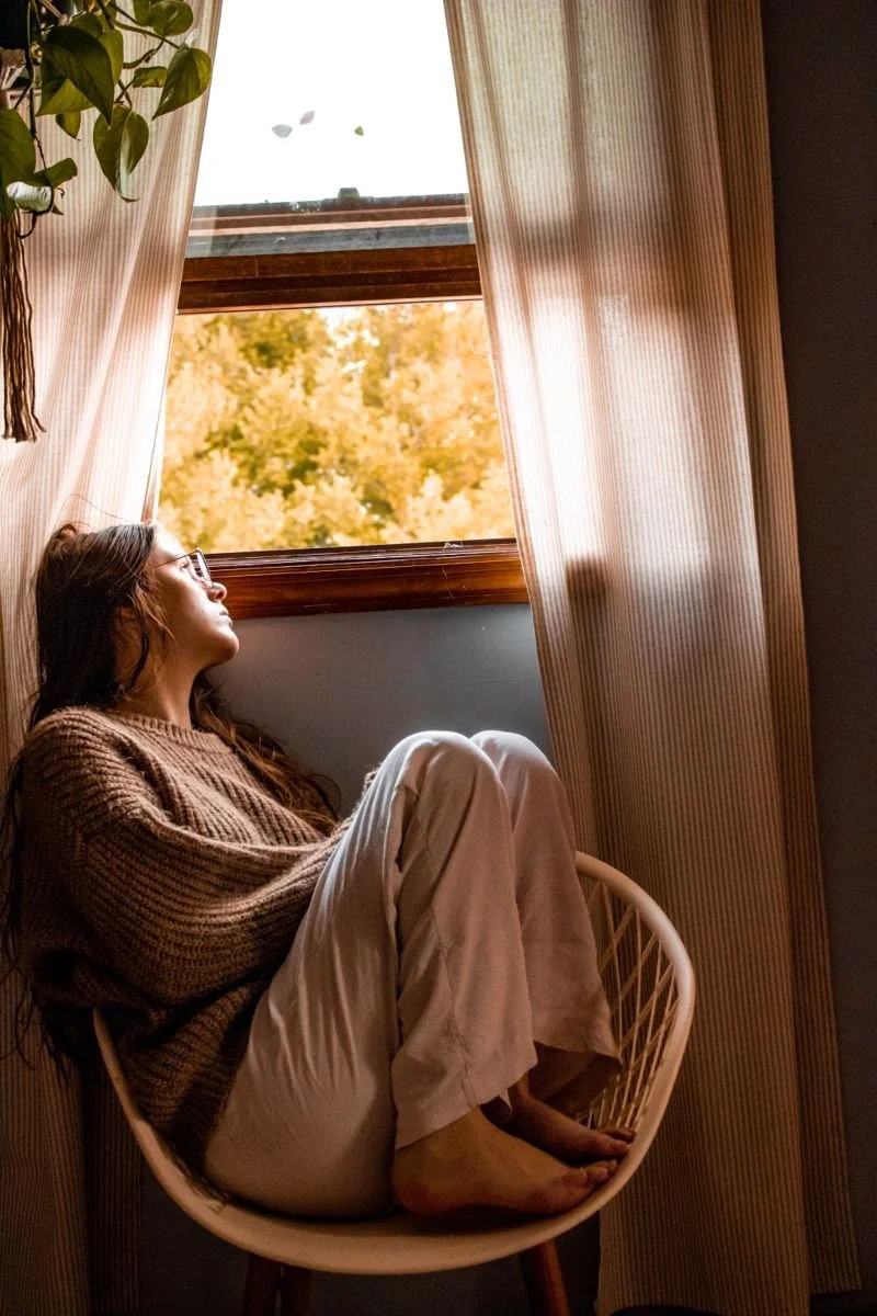 A woman with glasses and long hair sits curled up in a chair near a window with beige curtains, looking outside at fall-colored trees.