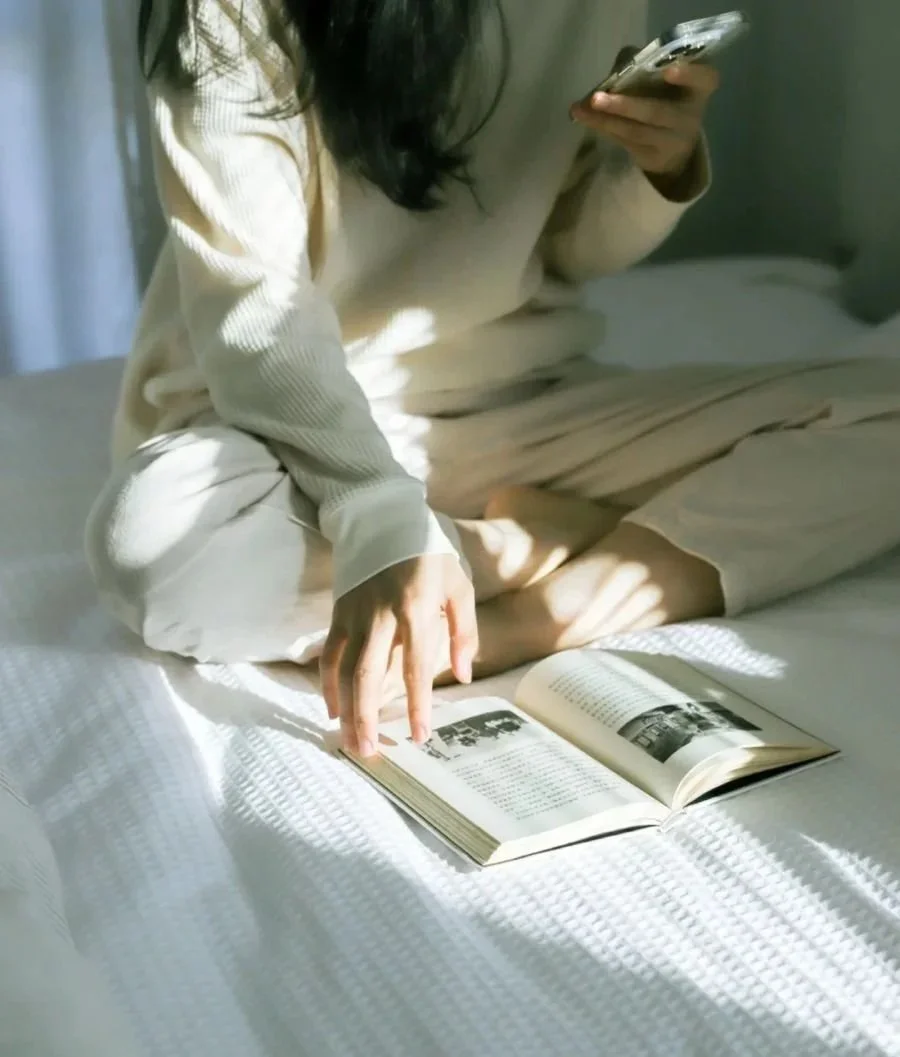 A person with long dark hair wearing cream-colored pajamas is sitting on a bed, reading a book with black and white photographs. Natural light creates shadows on the bed and the person's hand.