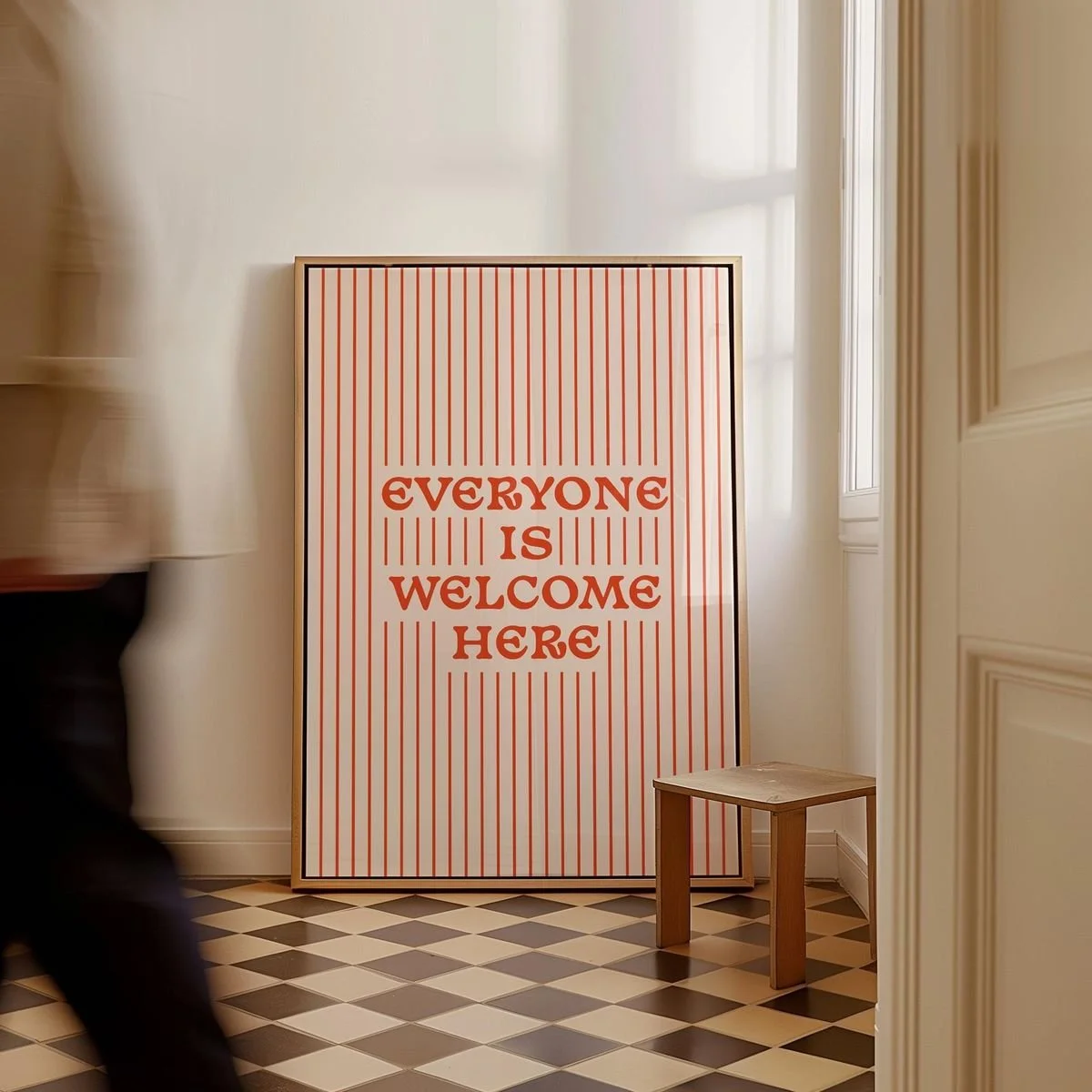 A white sign with orange vertical stripes and orange text that says, "Everyone is Welcome Here," leaning against the wall near a window and a small wooden table.