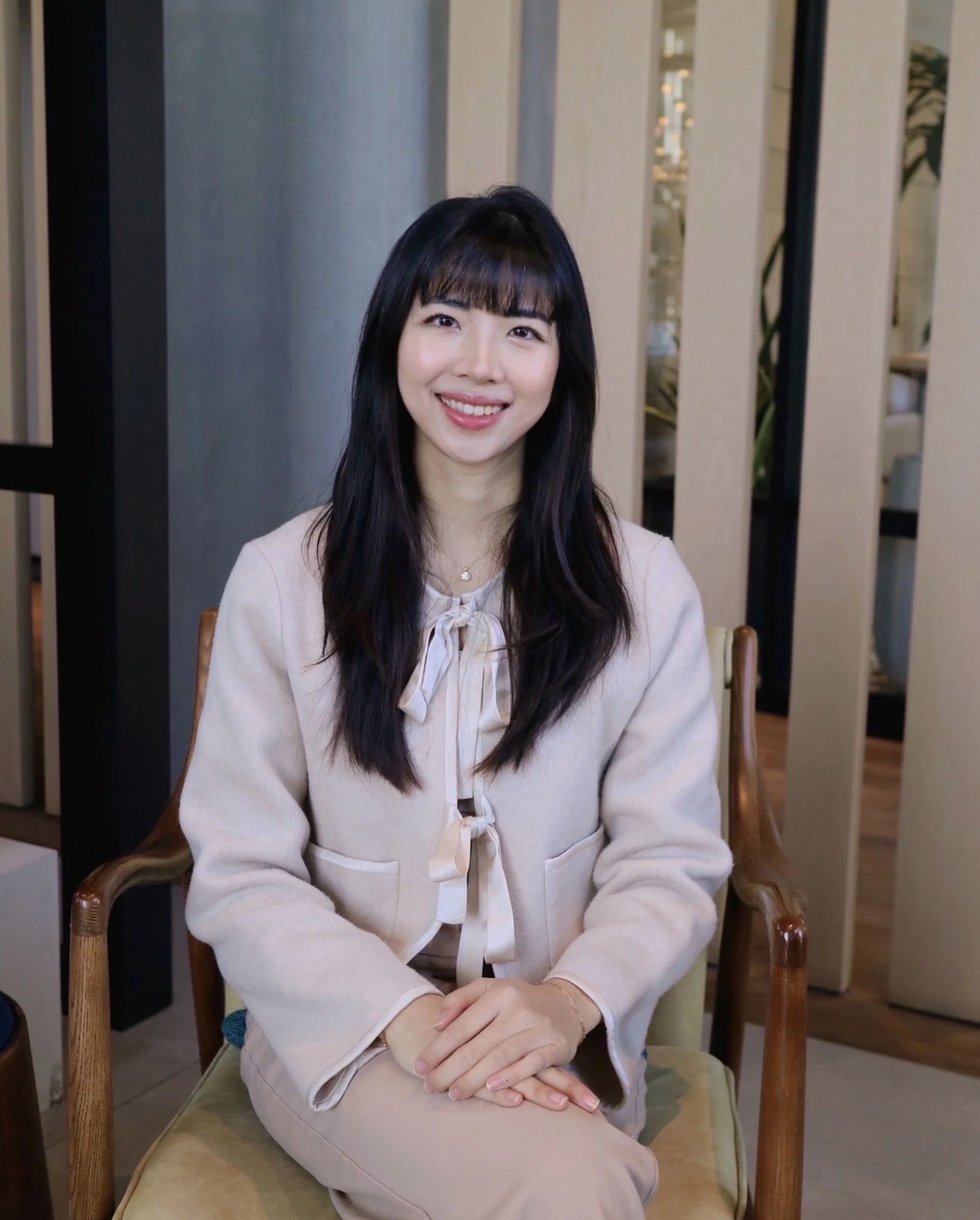 A woman with long dark hair and bangs smiling while sitting in an indoor setting.