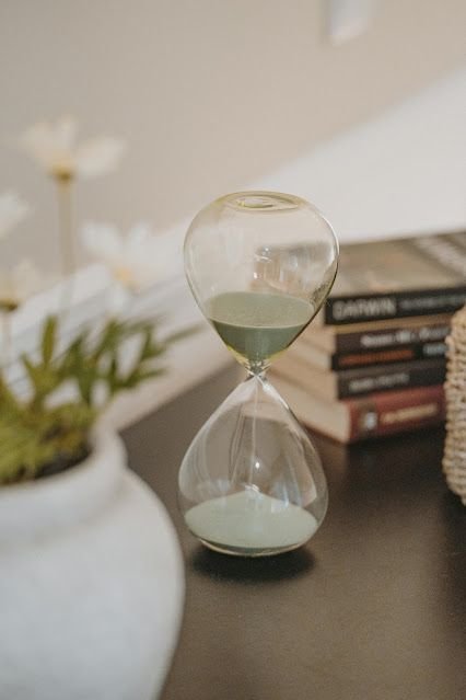 An hourglass with green sand on a black surface, with a white flower in a vase and a stack of books in the background.