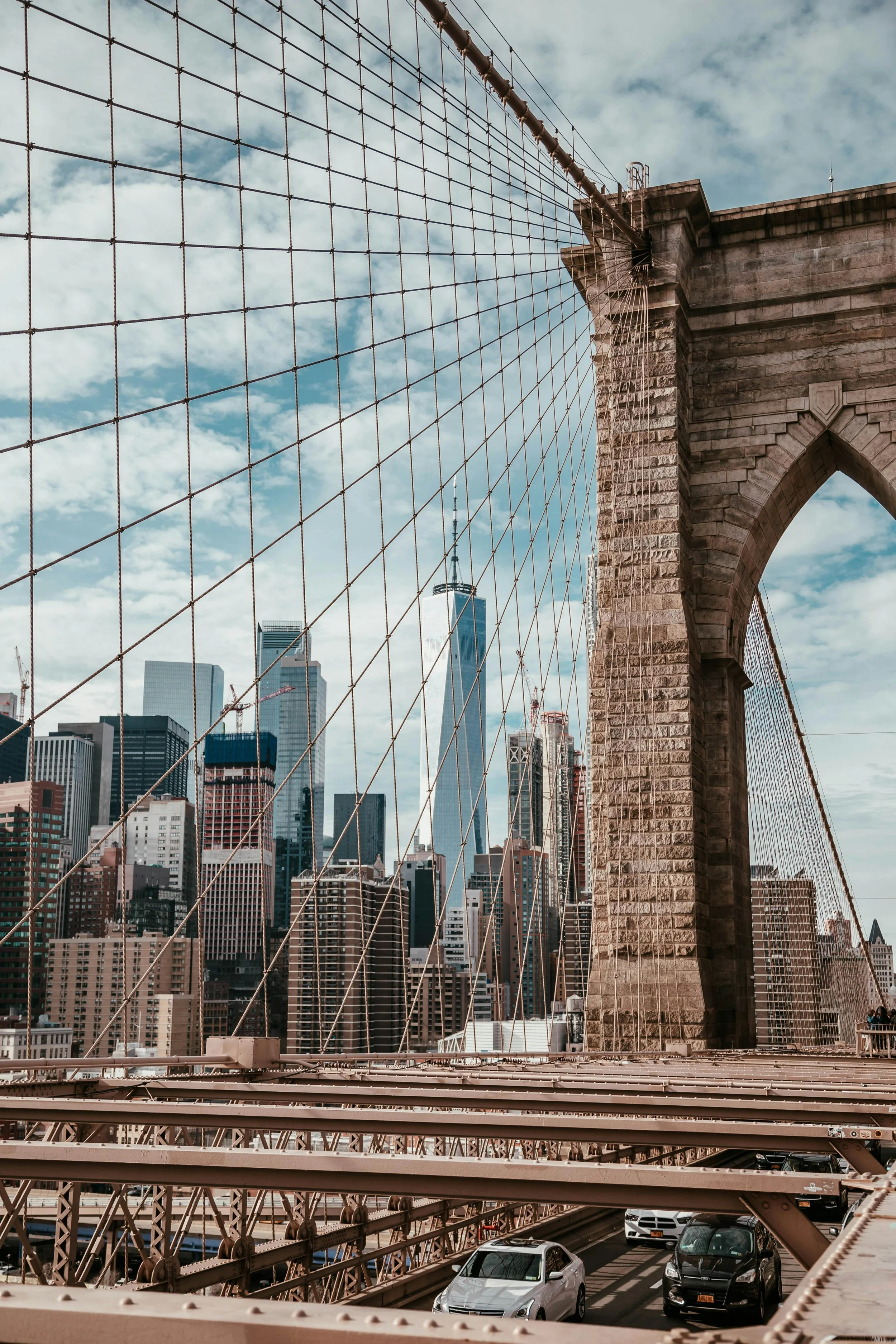 View of the Brooklyn Bridge with city skyline of Manhattan in the background, route of cars on the bridge, cloud-filled sky.