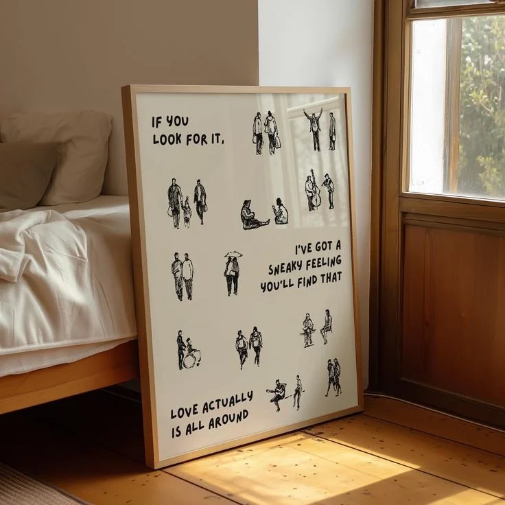 A large framed whiteboard with black illustrations of people in various poses, resting against a wall near a window; the board has text that reads, 'If you look for it, I've got a sneaky feeling you'll find that love actually is all around.'
