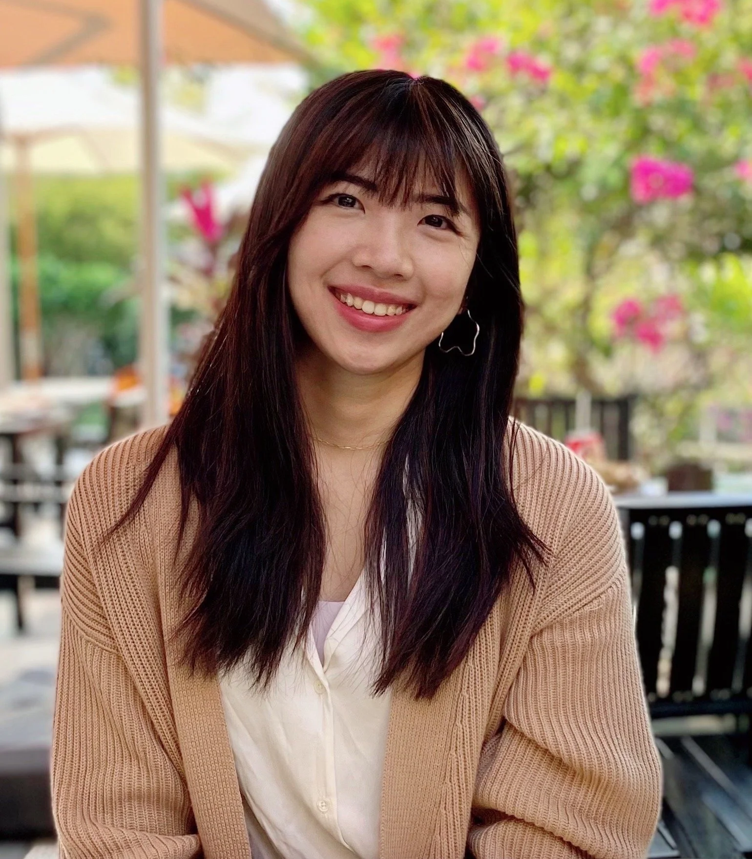 A woman with long dark hair and bangs, smiling, sitting outdoors at a restaurant or cafe with pink flowers and greenery in the background.